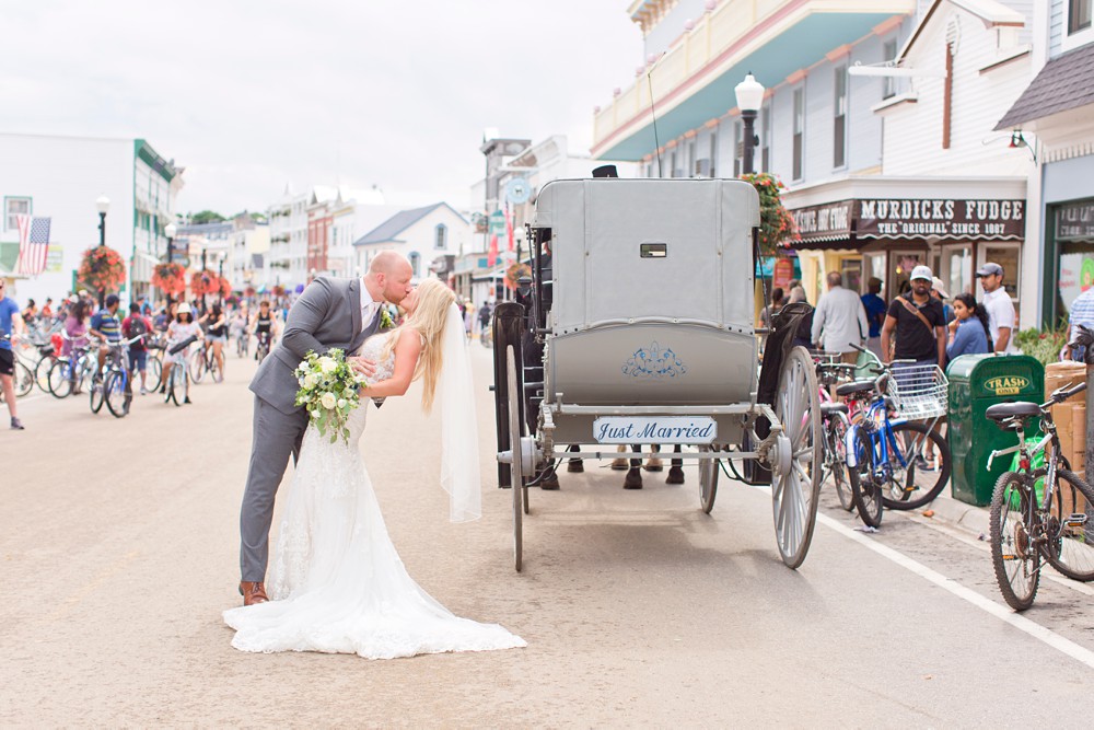 Bride and groom kissing next to horse drawn carriage in the middle of Main Street at destination wedding on Mackinac Island