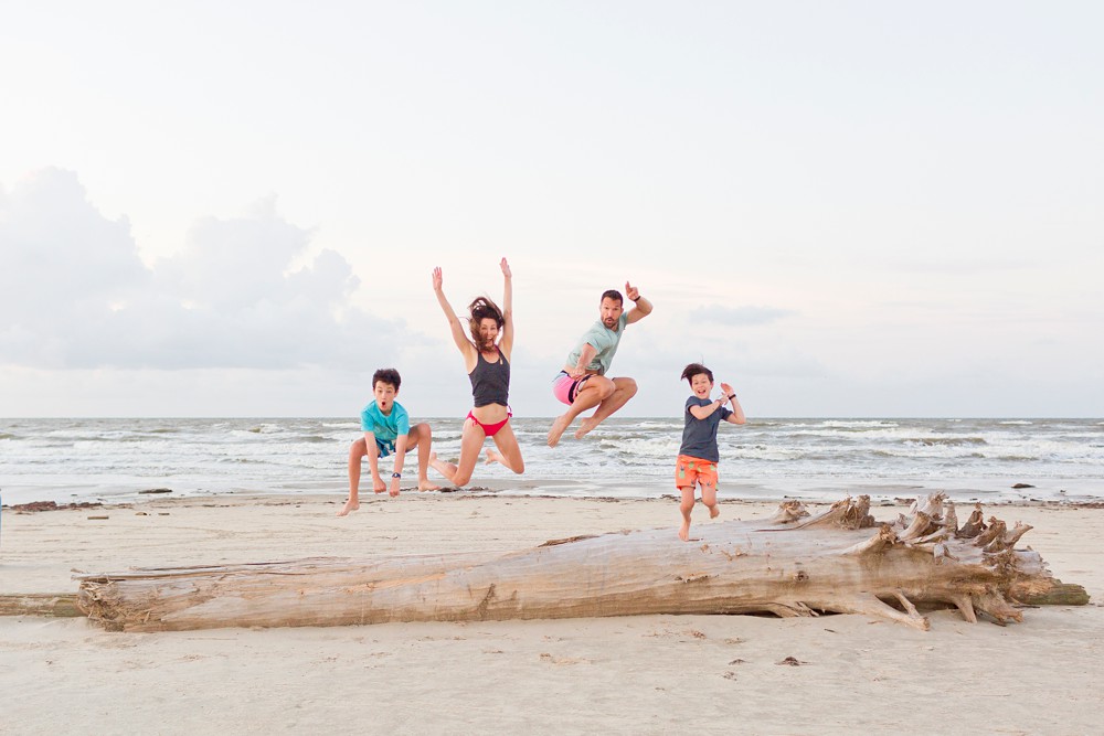 Family playfully jumping off a large piece of driftwood on a Galveston beach