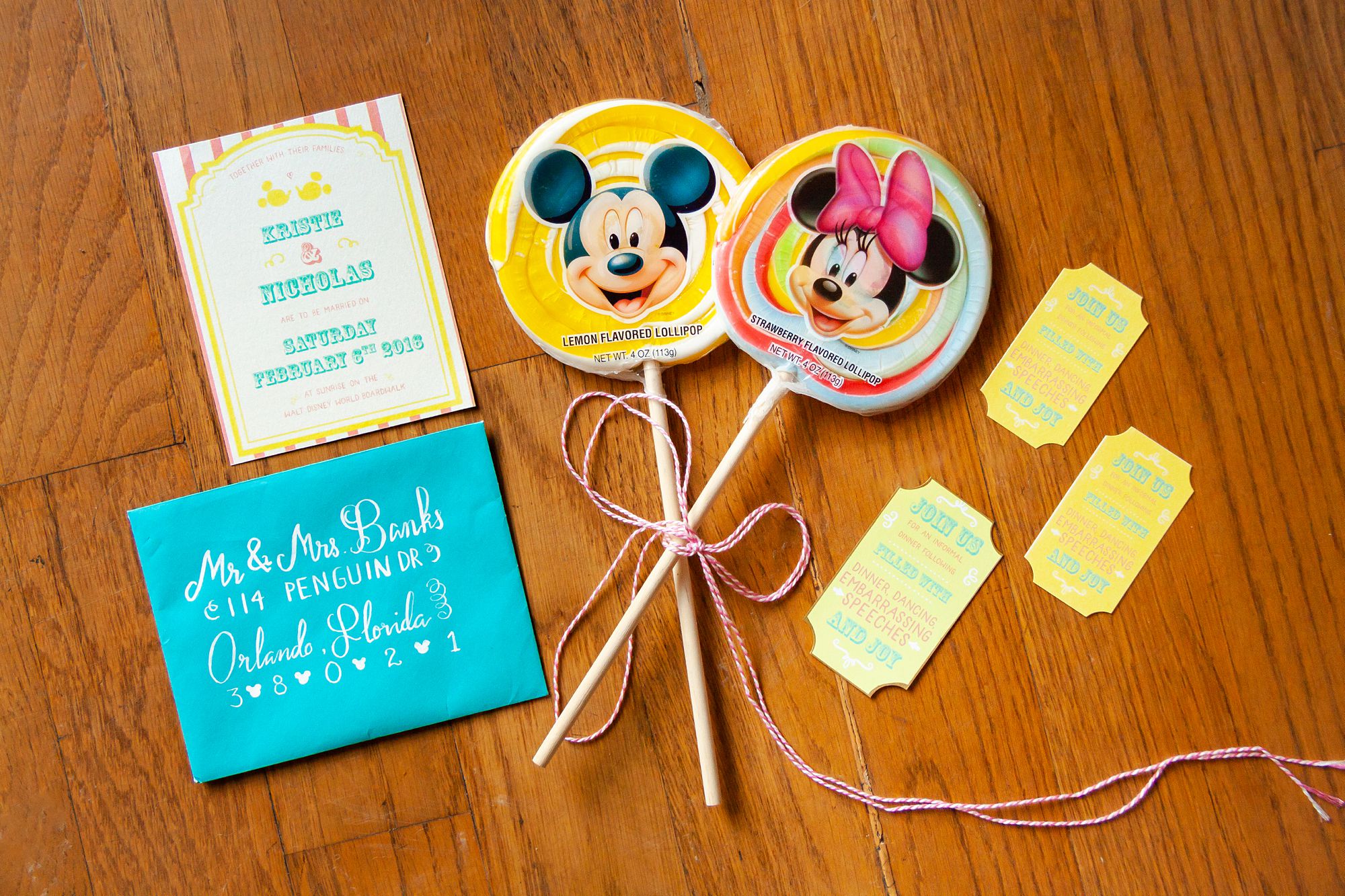 A pink, yellow and turquoise Disney carnival inspired wedding invitation and turquoise envelope is displayed with themed tickets and Mickey Mouse and Minnie Mouse lollipops.