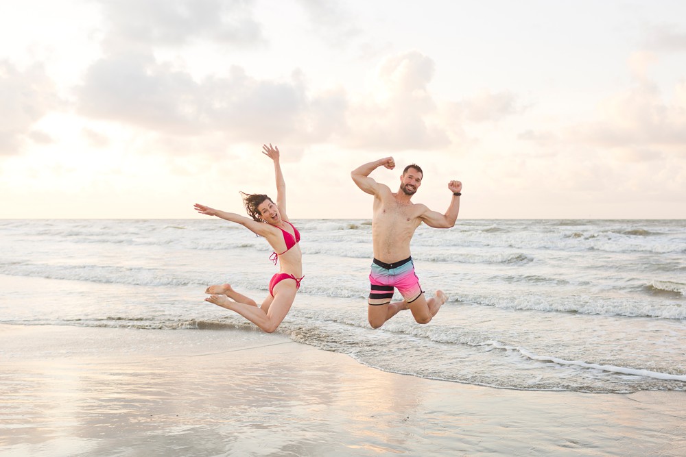 husband and wife jumping on the beach with the sunrise behind them