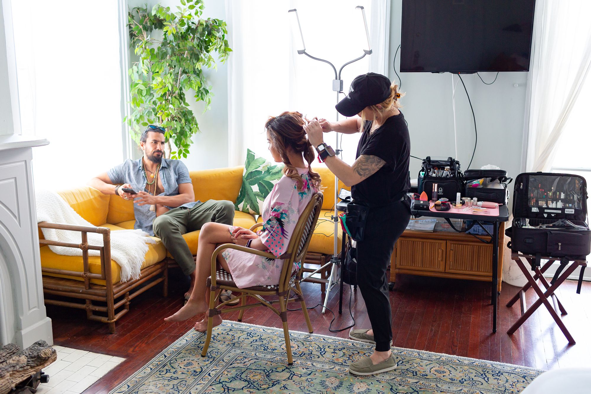 A groom sits on a couch while a hairstylist works on the a bride's hair.