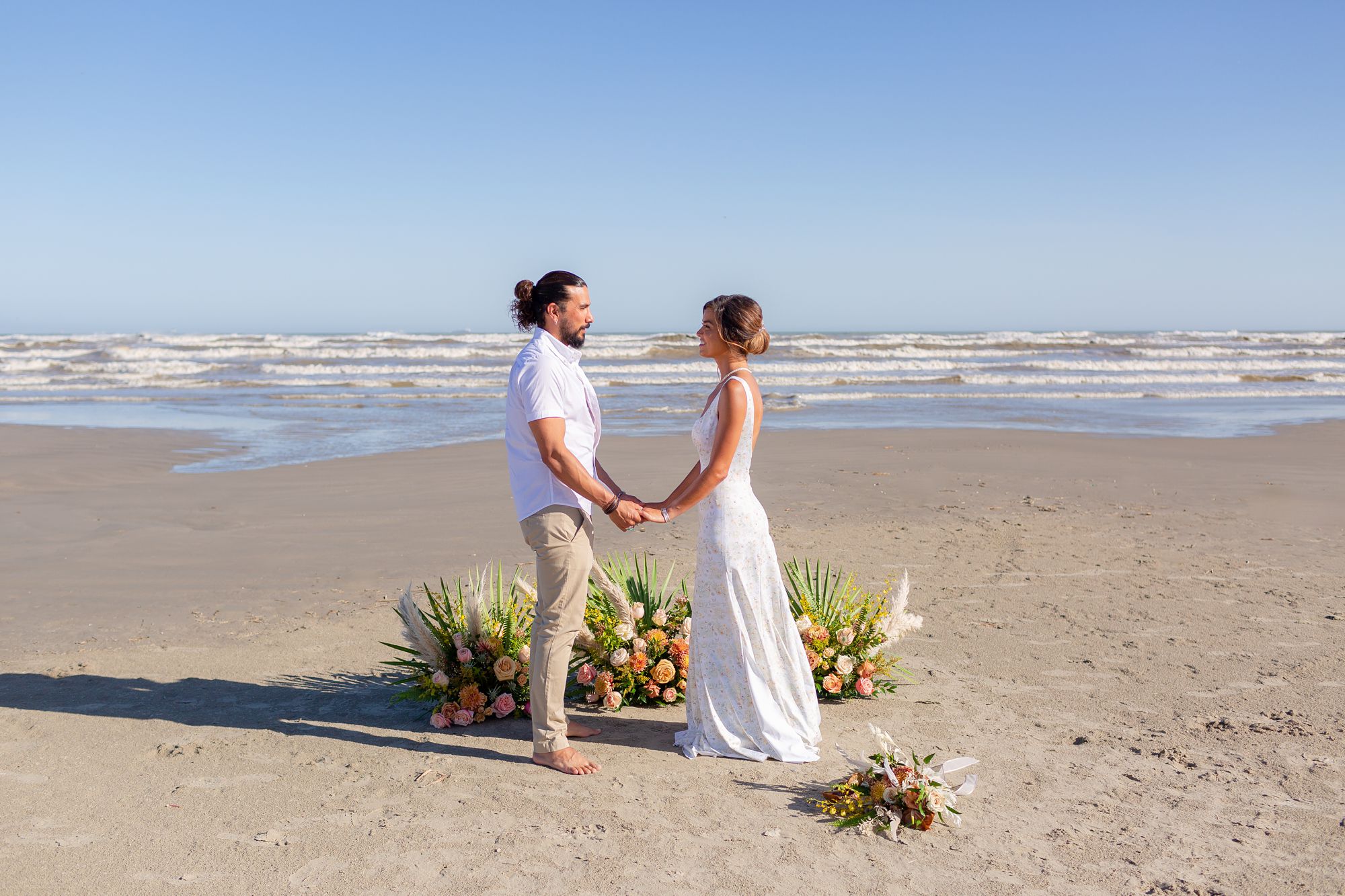 A bride and groom stand facing each other and holding hands in front of a ceremony flower arrangement at their Preserve at Grand Beach elopement.