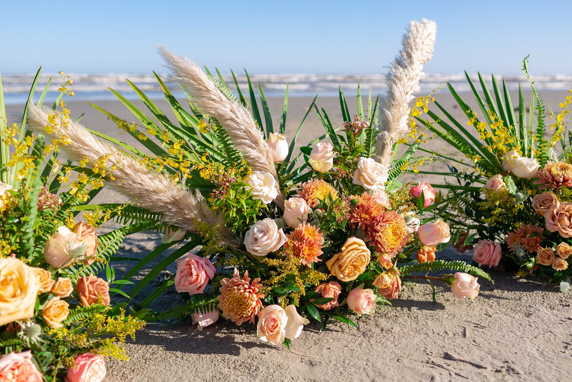 Three large flower arrangements with pink and orange roses and pampas grass sit in the sand at a Preserve at Grand Beach elopement in Galveston.