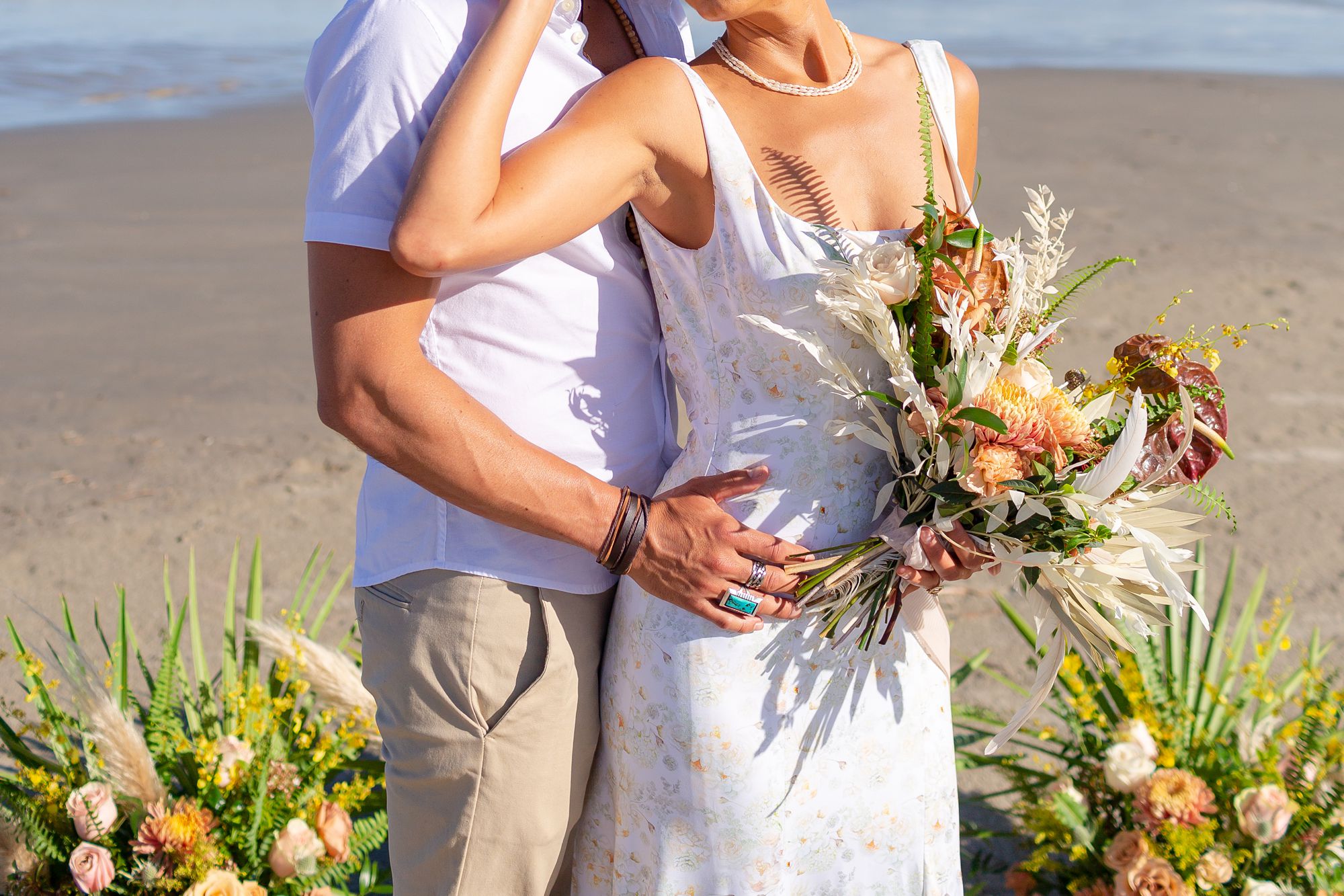 A groom holds bride around the waist in front of their elopement ceremony flower arrangements.