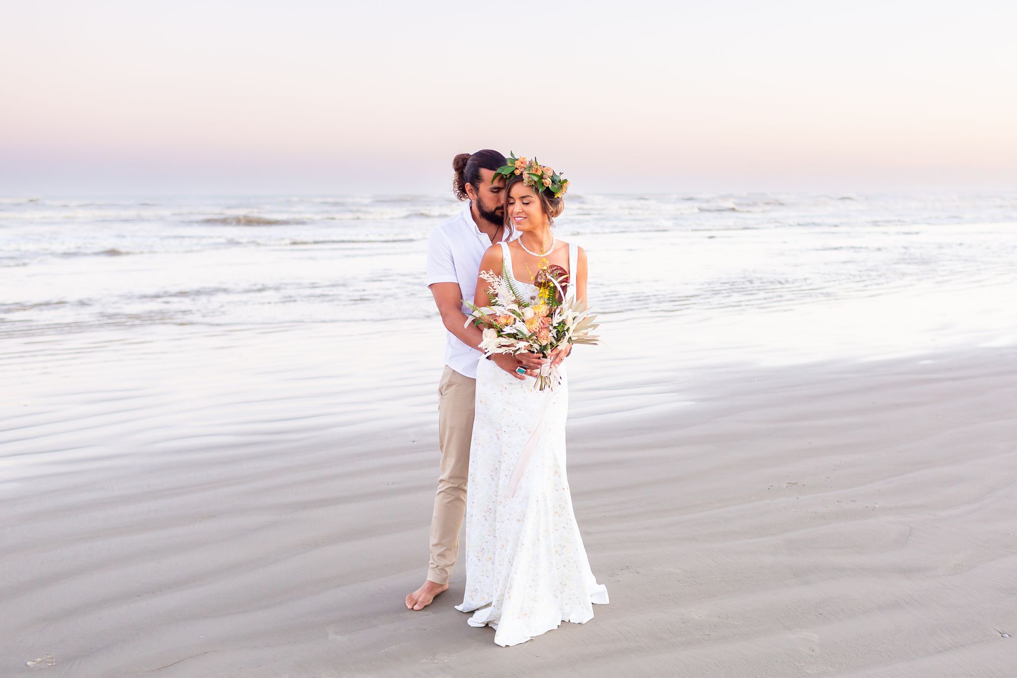 A groom holds his bride around the waist on the beach in Galveston at sunset.