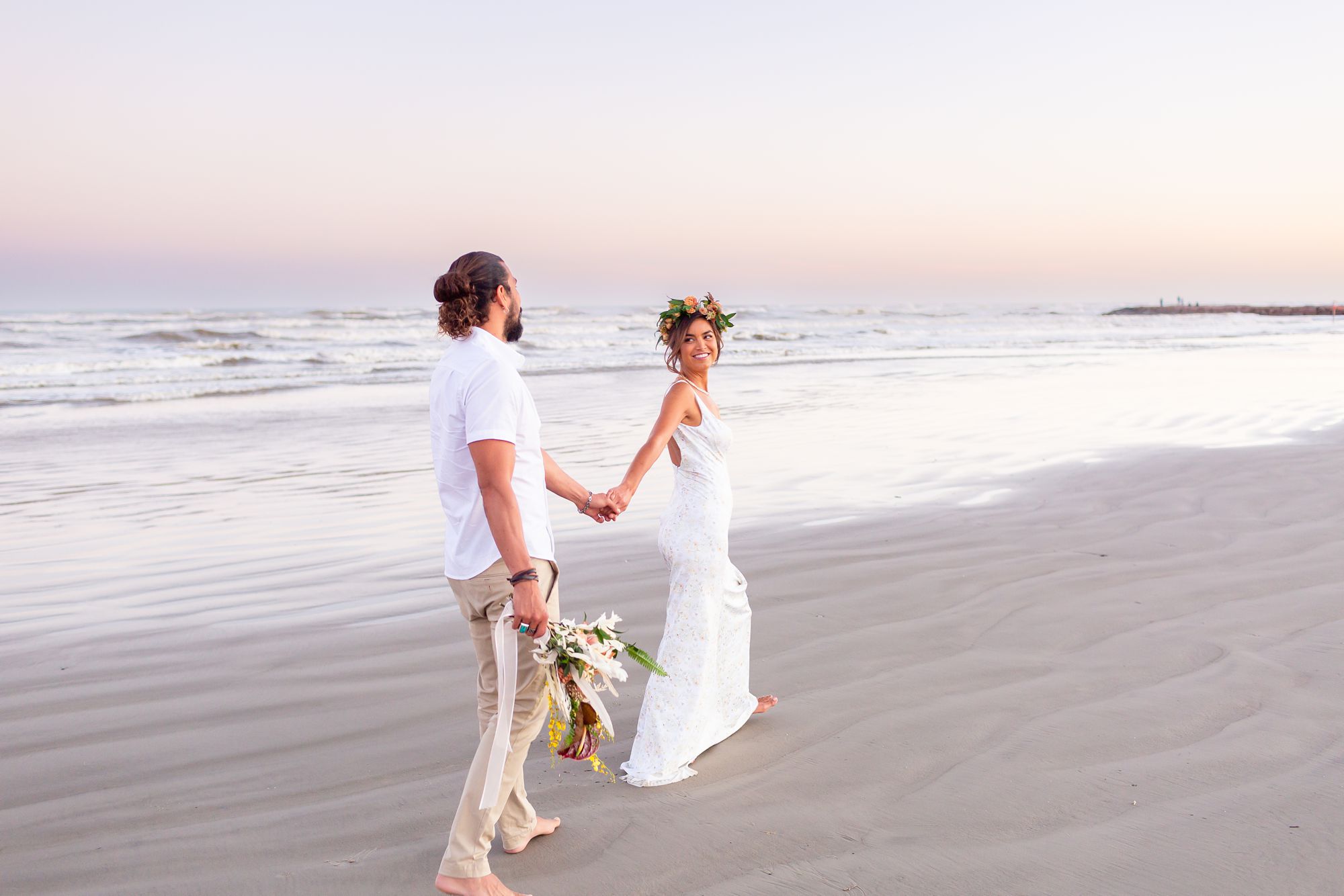 A bride and groom walk along the beach in Galveston at sunset.