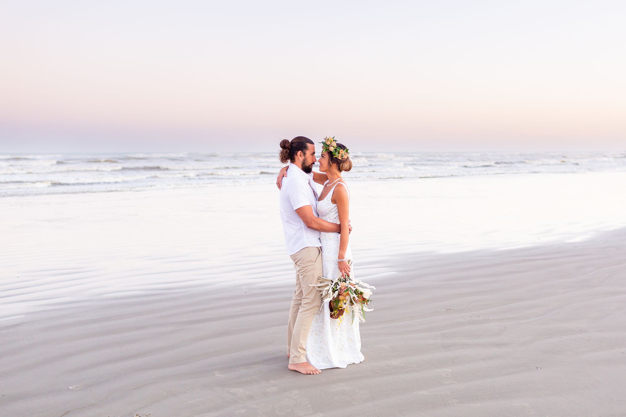 A bride and groom embrace on the beach and smile at each other with the ocean and a pink sky sunset in the background.