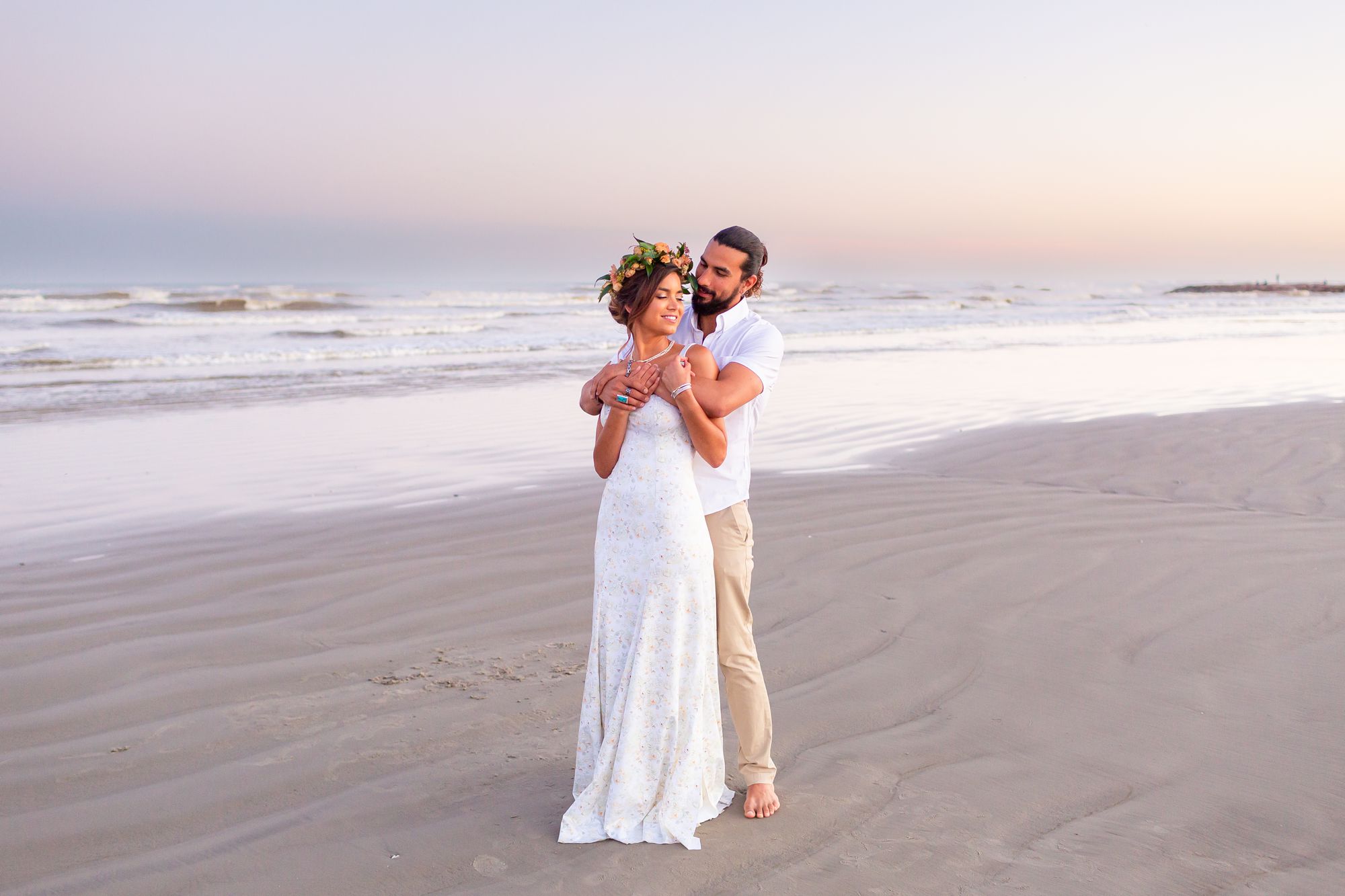 A groom holds his bride around the shoulders on the beach in Galveston at sunset.