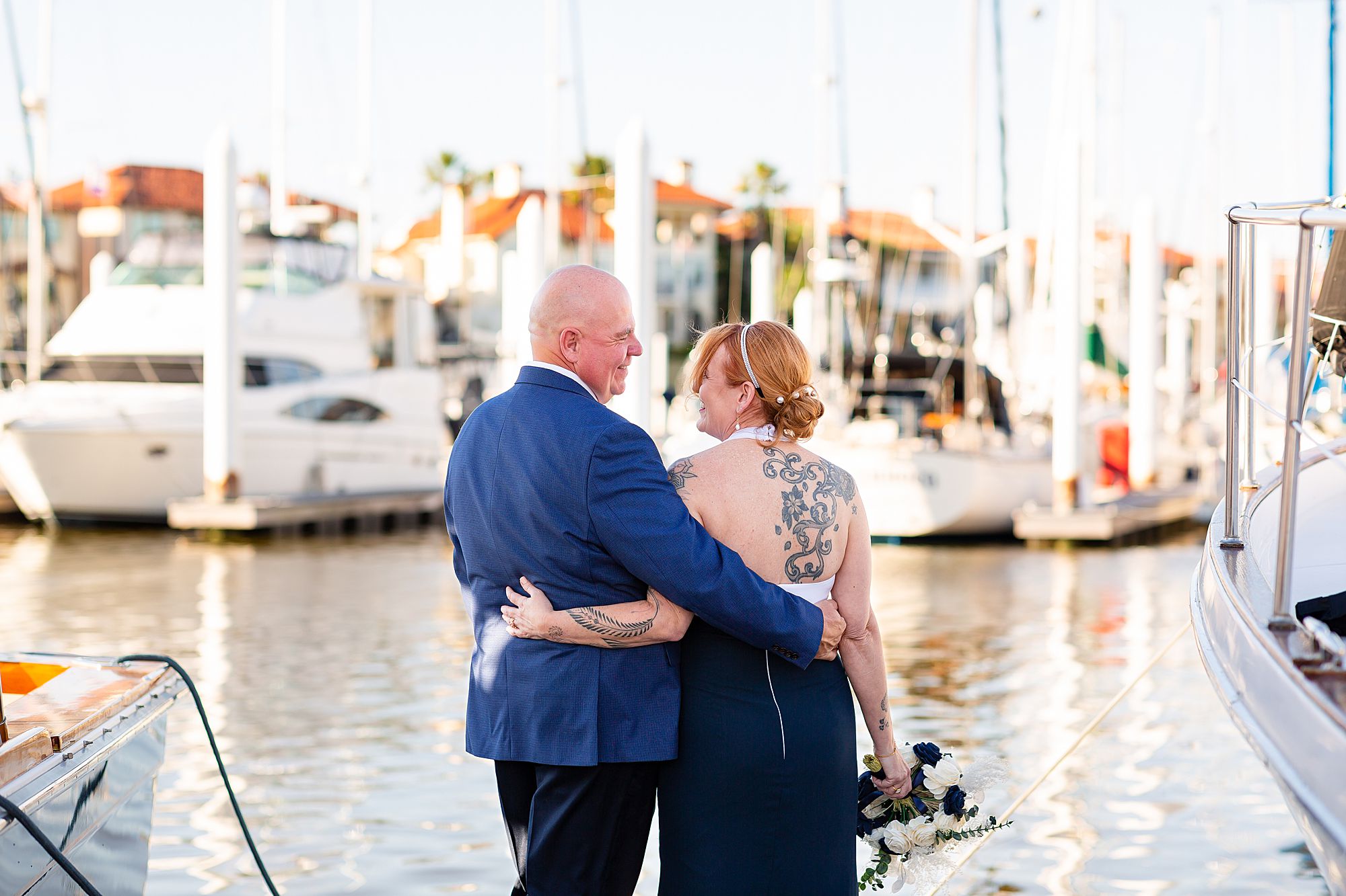 A bride and groom stand on a dock with their arms around each other and smile at each other.