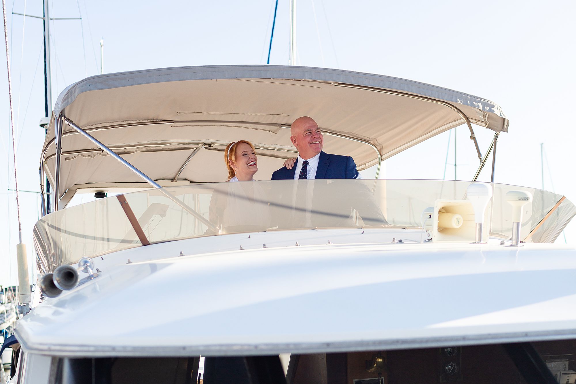 A bride and groom stand on the fly bridge of a motor yacht looking out at the water.