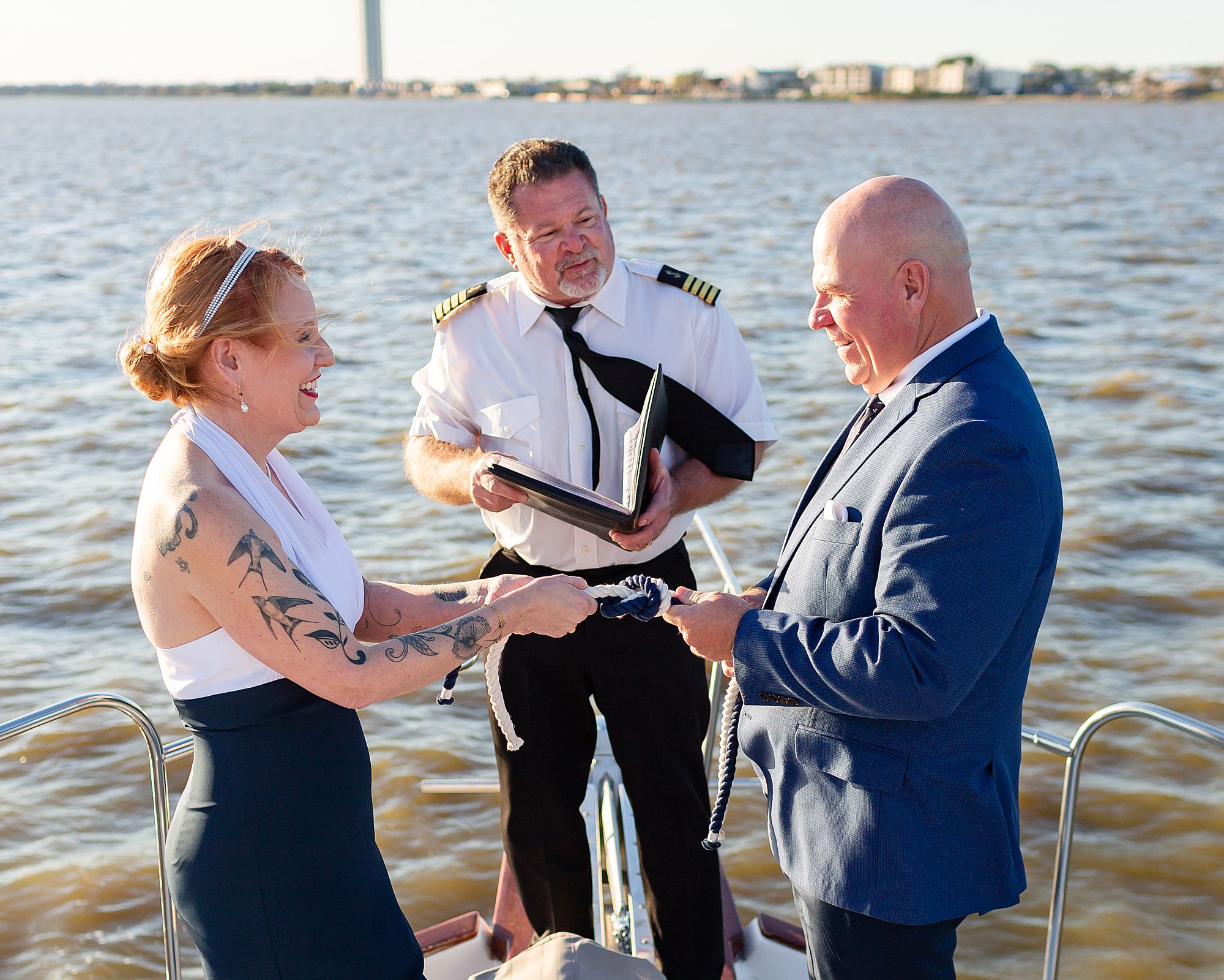 A bride and groom standing on the bow of a yacht, gaze at each other as they pull a fisherman's knot tight during their elopement ceremony.