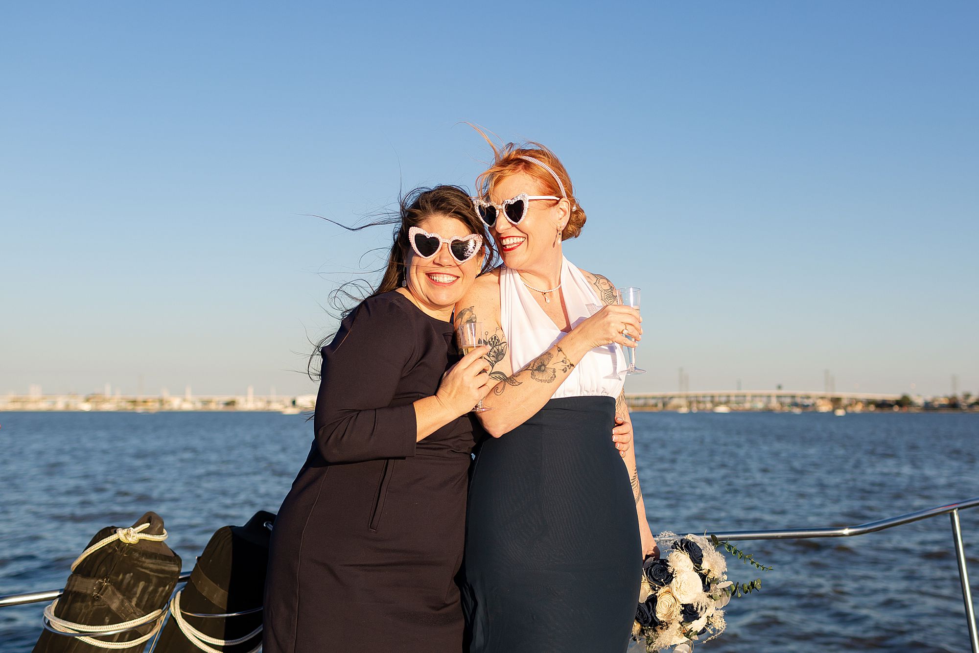 A bride and elopement guest wearing heart shaped sunglasses laugh with each other on a yacht in Kemah, Texas.