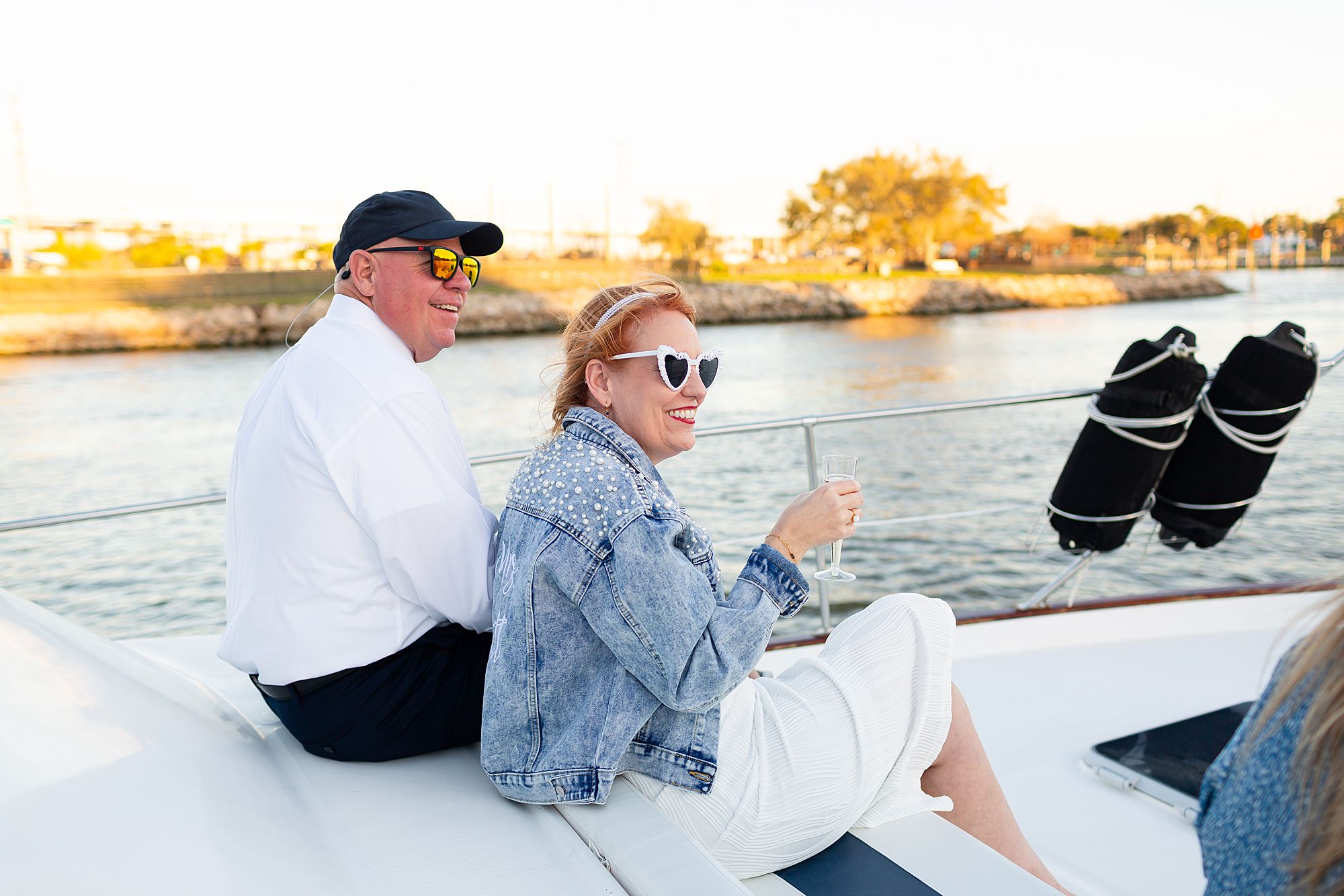 A bride and groom sit on a yacht drinking champagne at sunset.