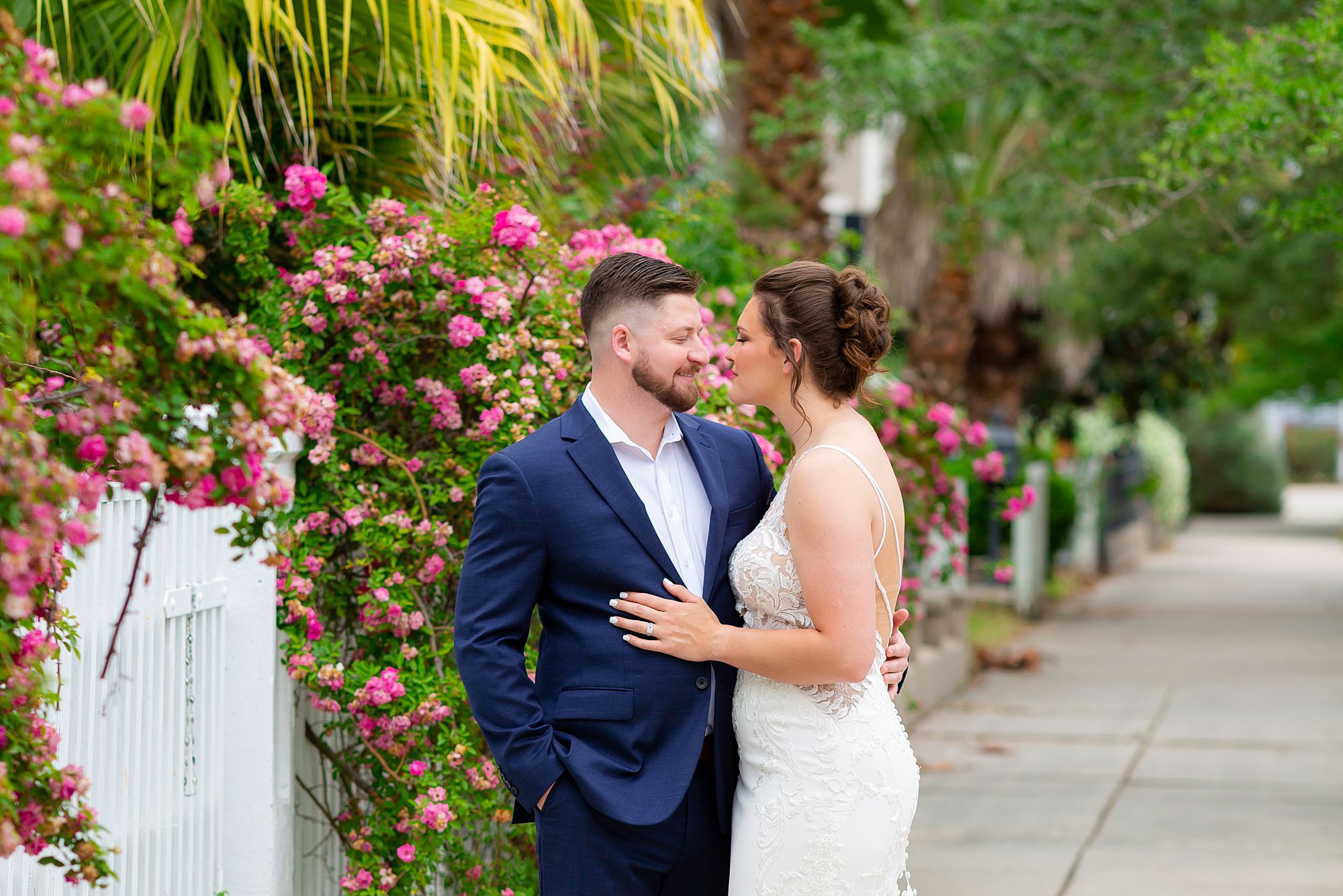 A bride and groom stand on a sidewalk in the east end historic district with flowers spilling over a white picket fence.