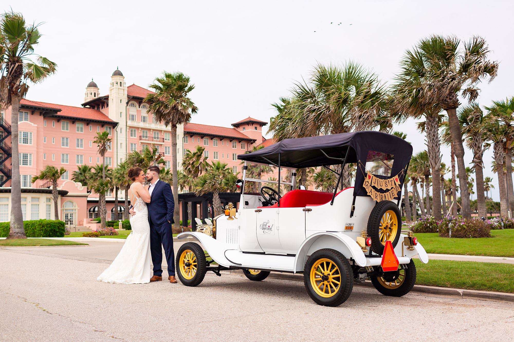 A bride and groom stand next to a Model T style golf cart in front of an early 1900s pink hotel on Galveston Island.