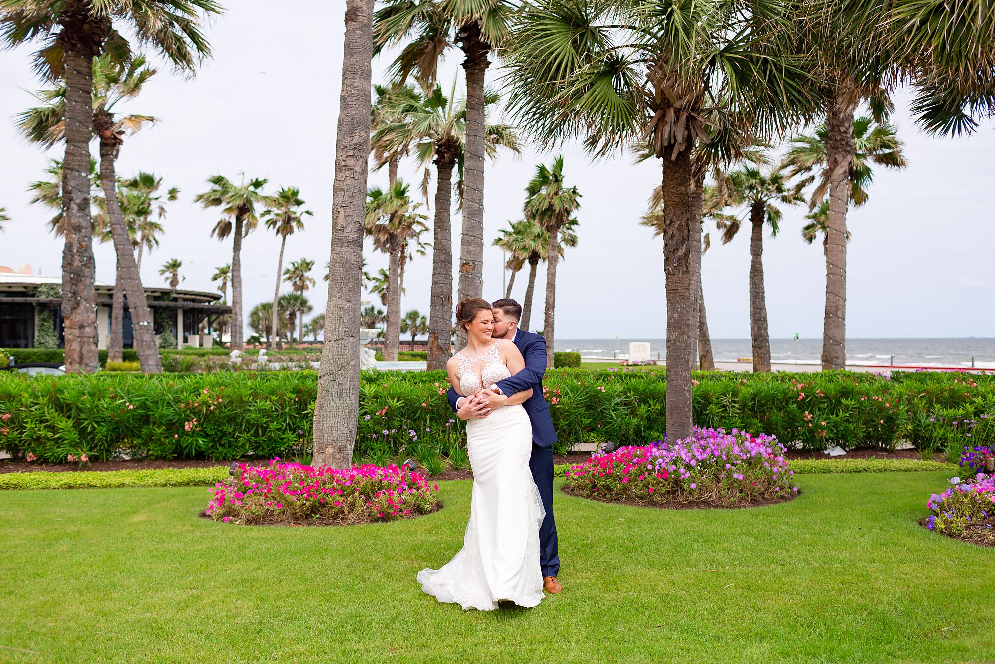 A bride and groom stand on a green lawn with palm trees and the ocean behind them.