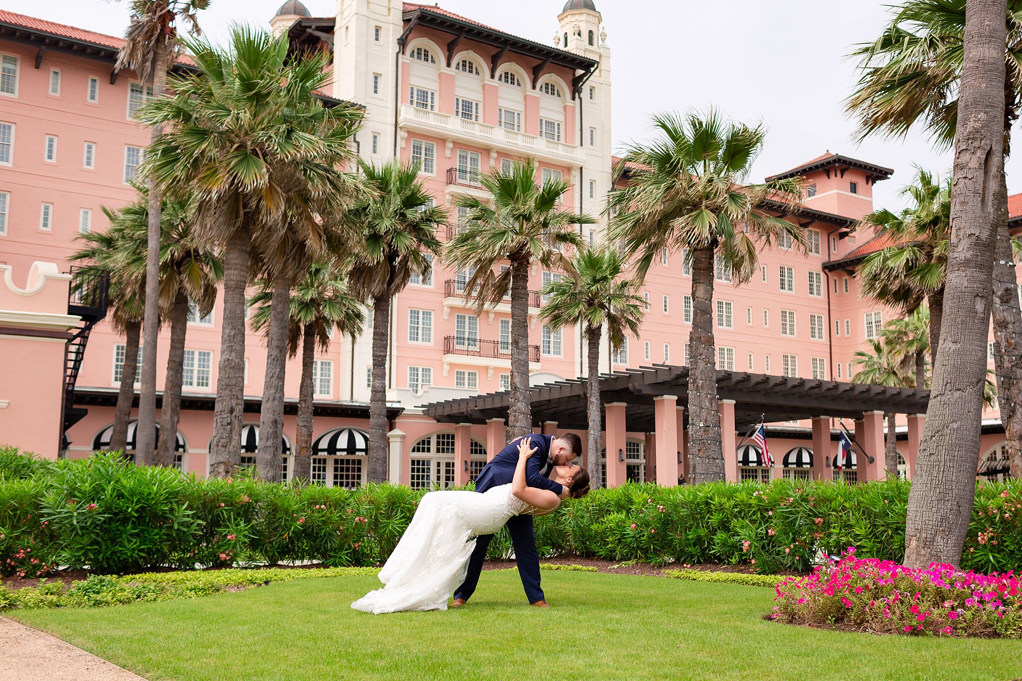 A groom dips his bride on the lawn of The Grand Galvez with the pink hotel and palm trees behind them.