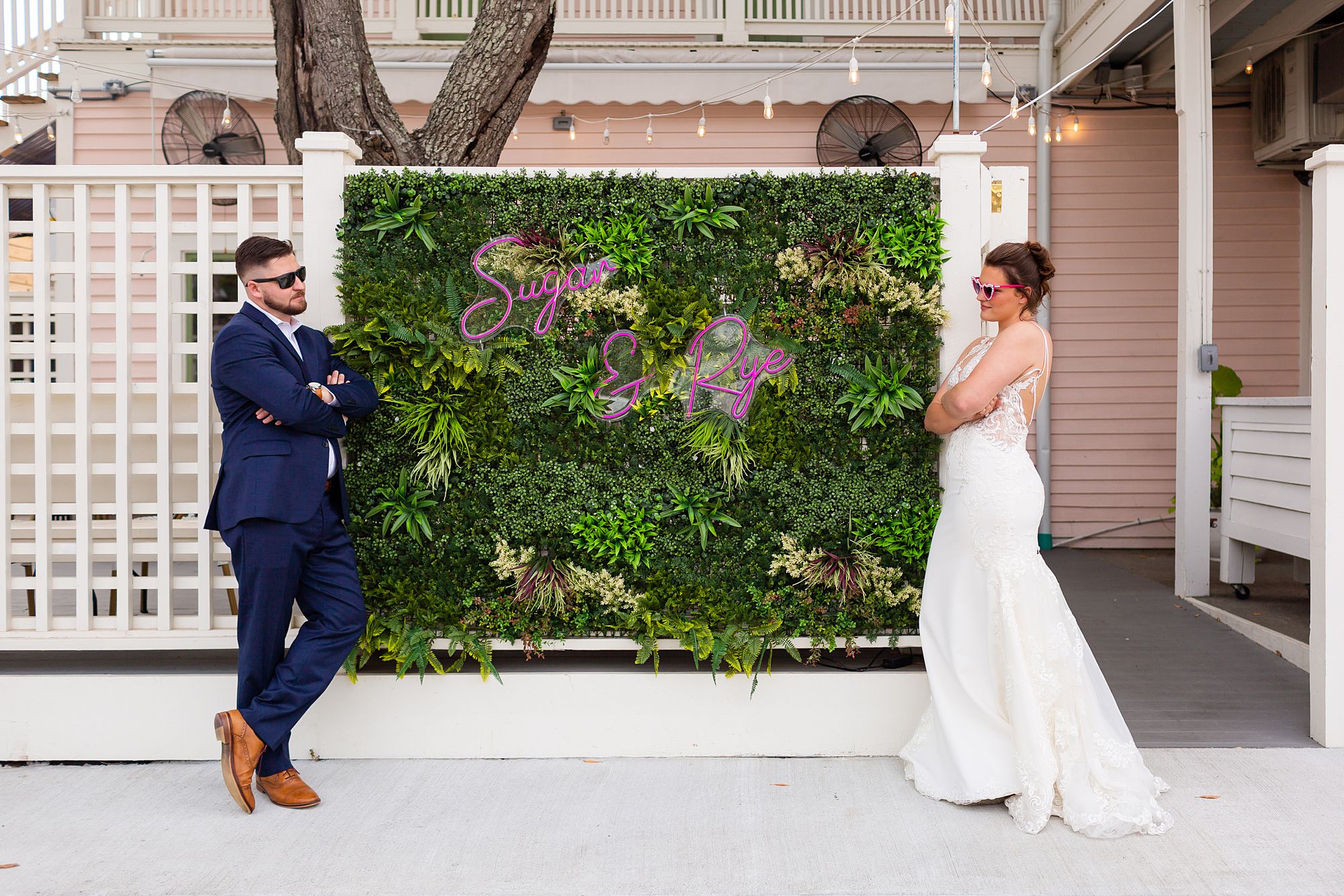 A bride and groom wearing sunglasses playfully look at each other in front of Sugar and Rye.