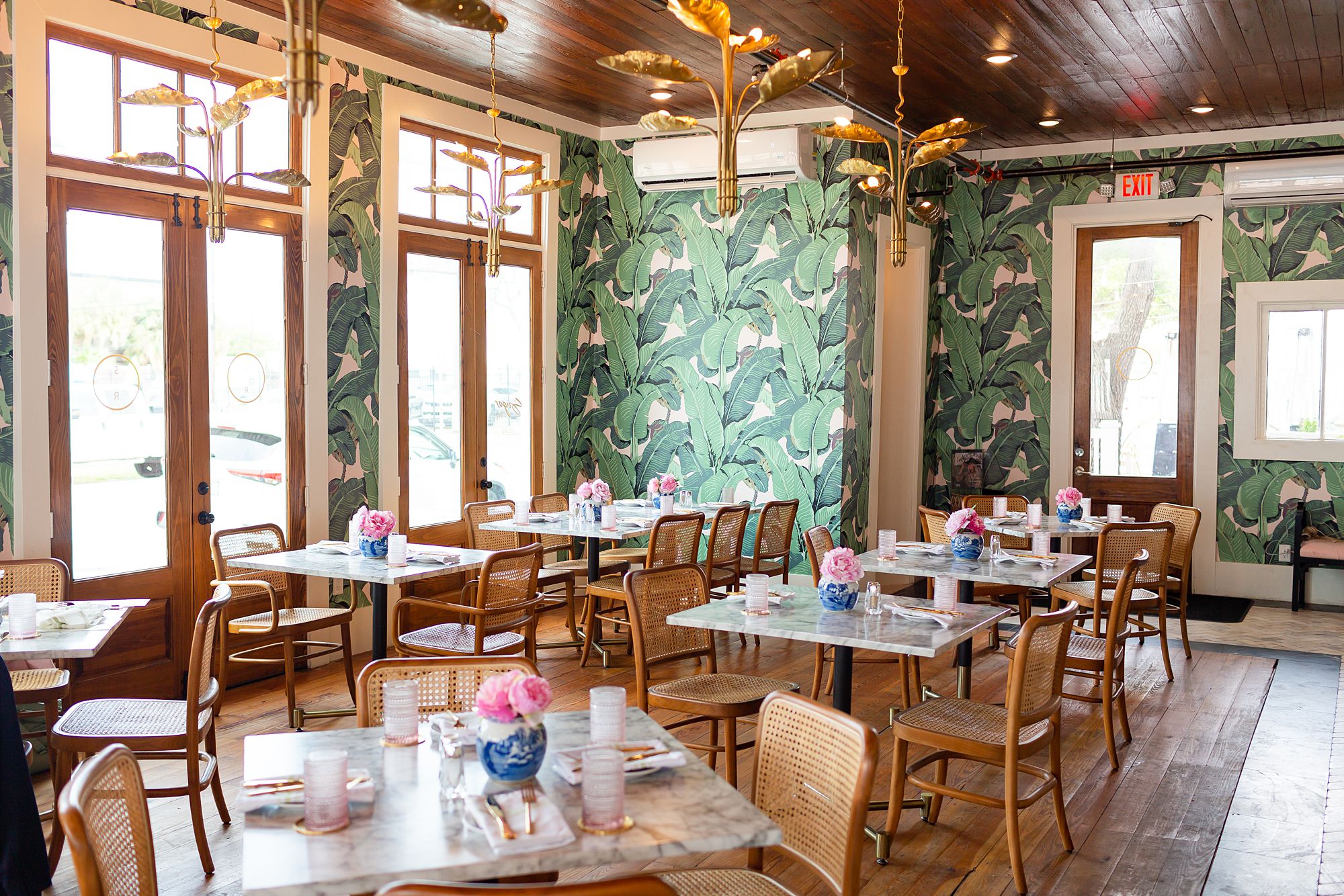 Sugar and Rye, a restaurant with pink and green palm frond wallpaper, marble top tables, and wicker chairs.
