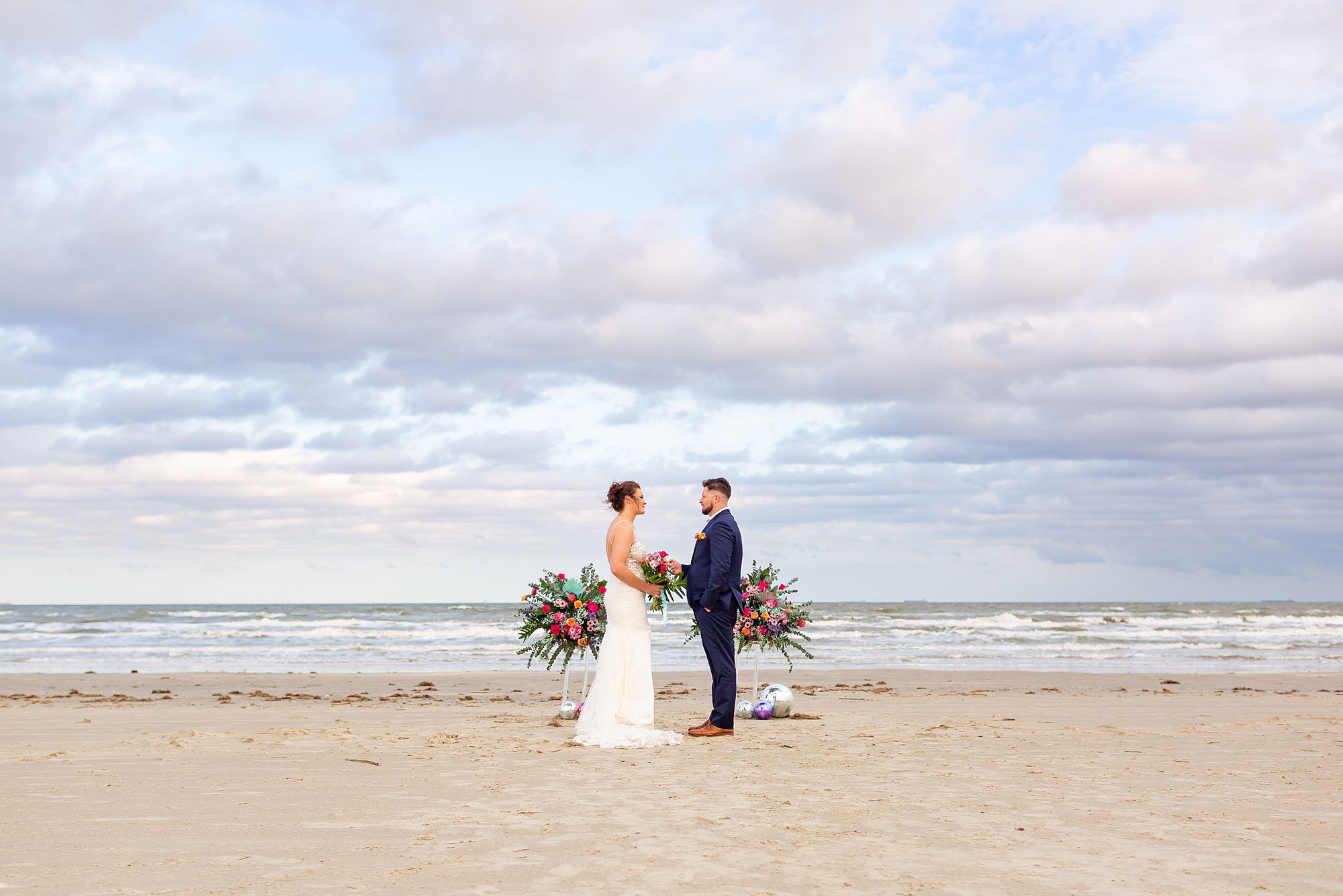 A bride and groom stand on a Galveston beach in front of a floral display sharing a vow renewal.