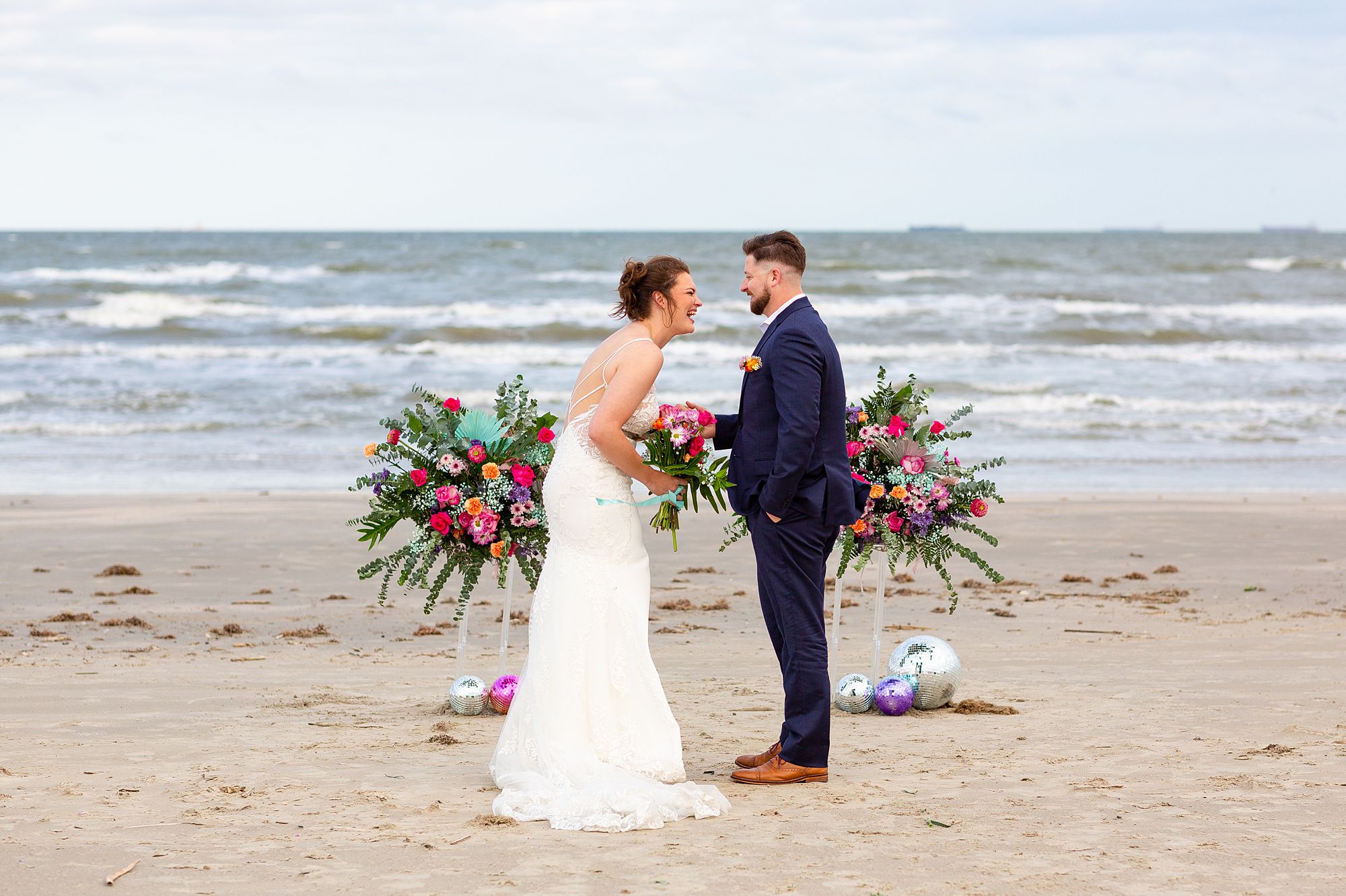 A bride and groom share a laugh on the beach in front of a floral display during a vow renewal at their elopement style anniversary.