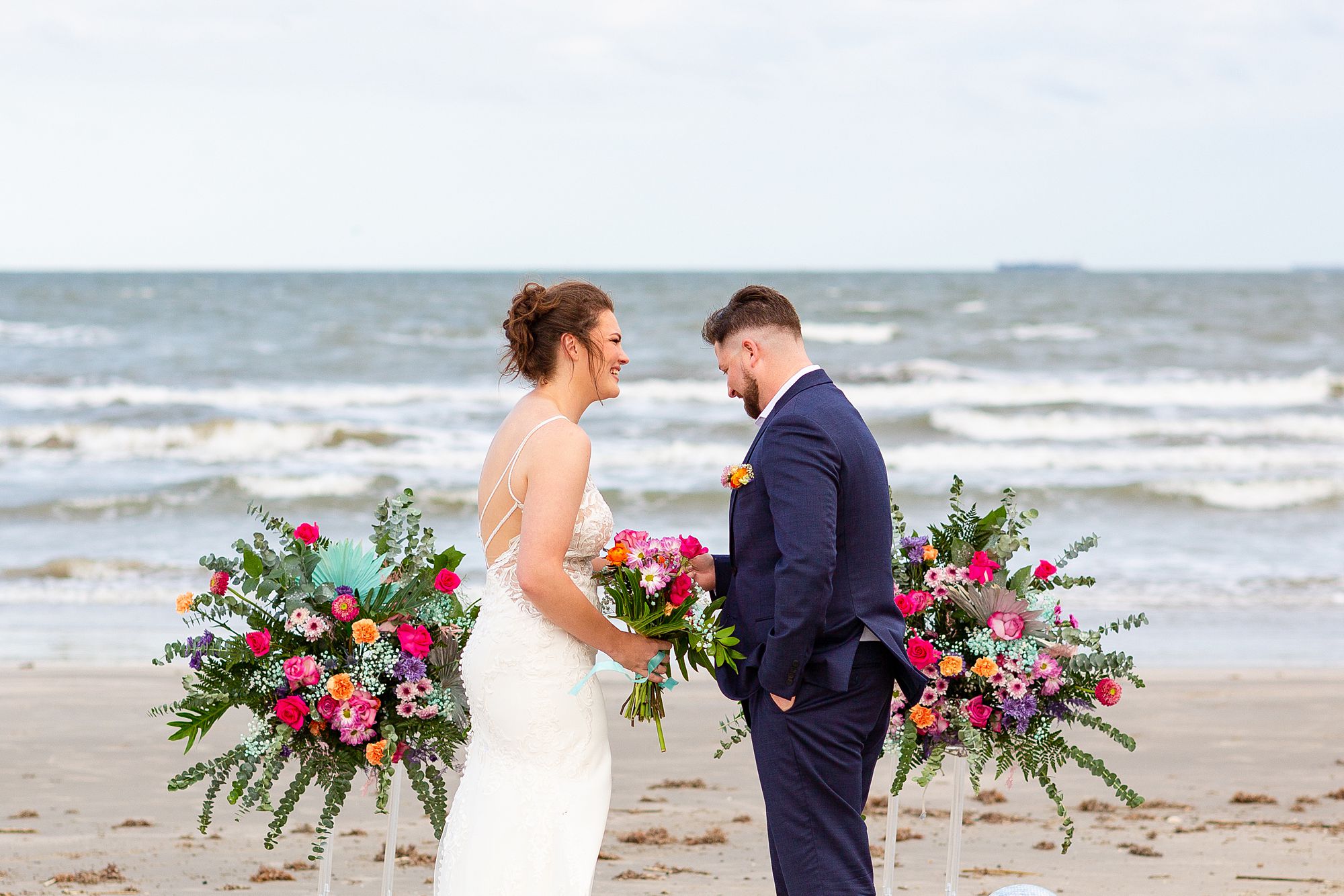 A bride and groom share a laugh on the beach in front of a pink, purple, and turquoise floral display during a vow renewal on Galveston Island.