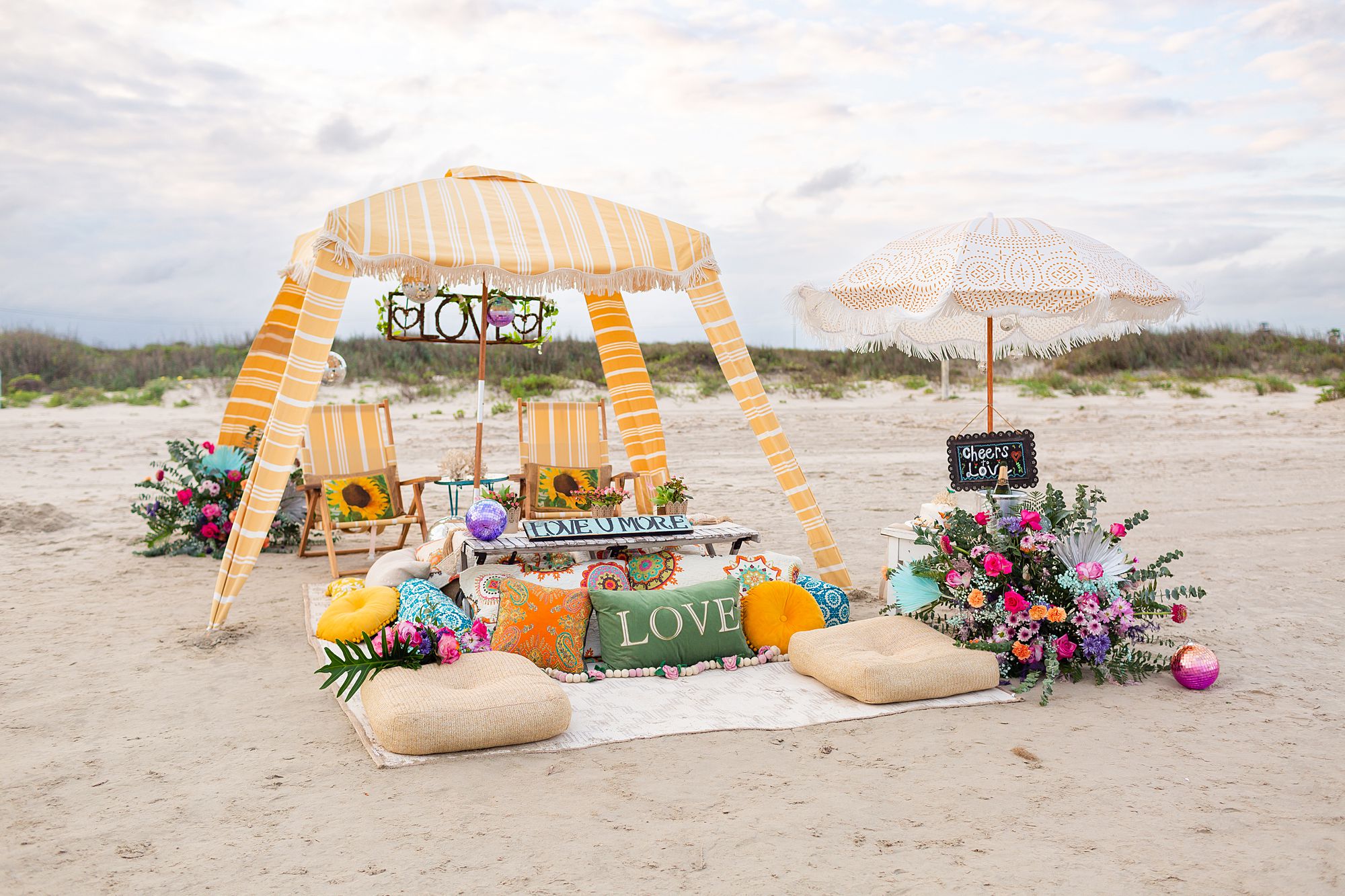 An elaborate beach picnic with a yellow canopy, beach chairs, cake and champagne table, rugs, and pillows.