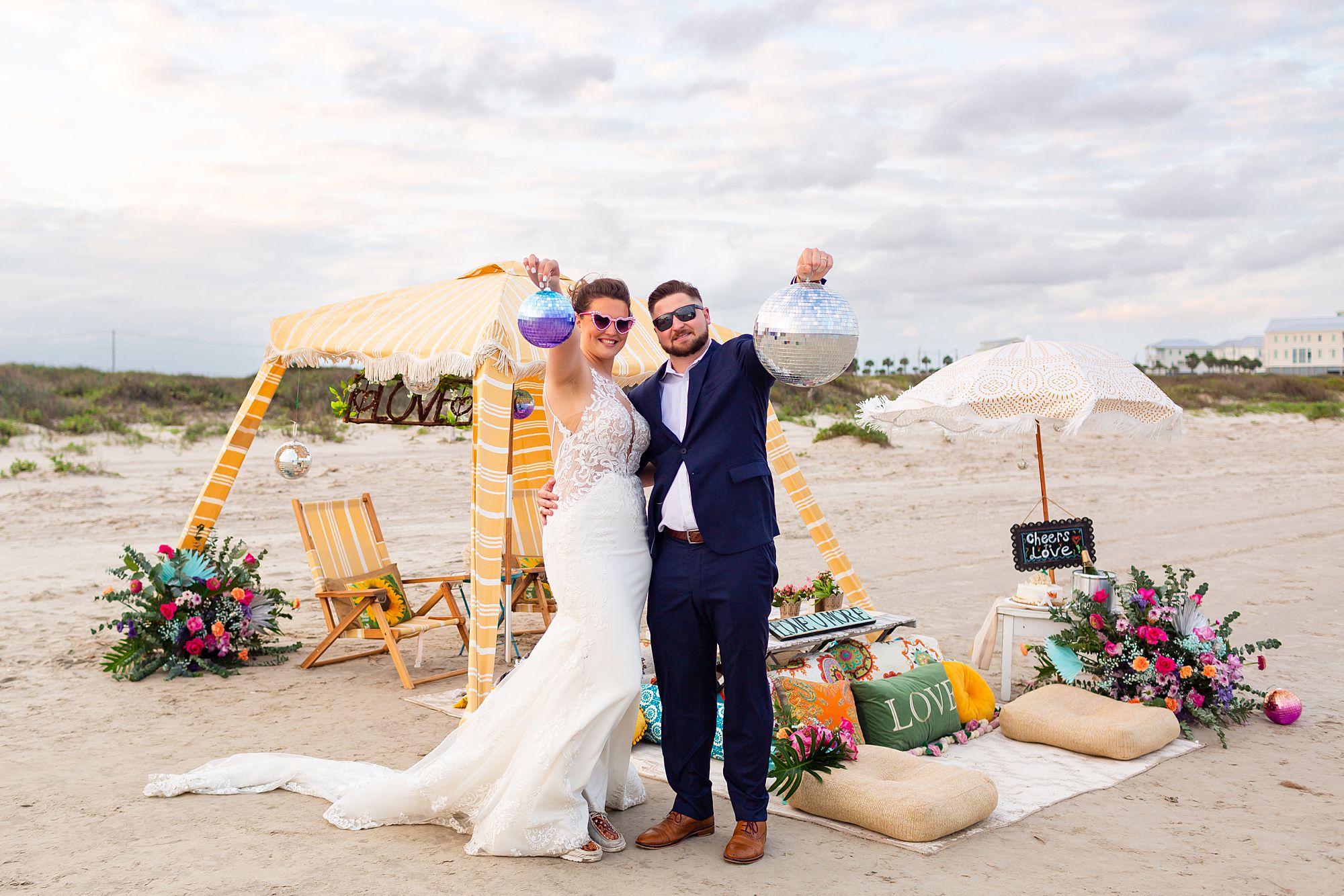 A bride and groom stand in front of a luxury beach picnic wearing sunglasses and holding up disco balls at their elopement style anniversary in Galveston.