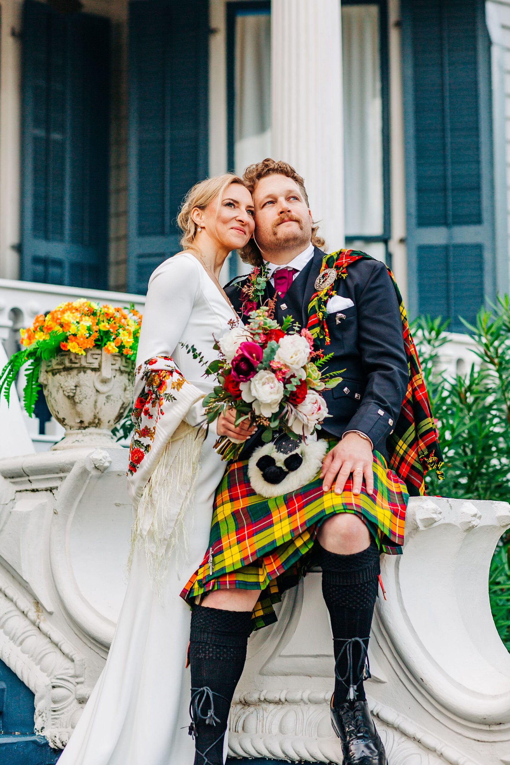 A bride in a white dress and a groom in a kilt stand together on the front stairs of Carr Mansion.