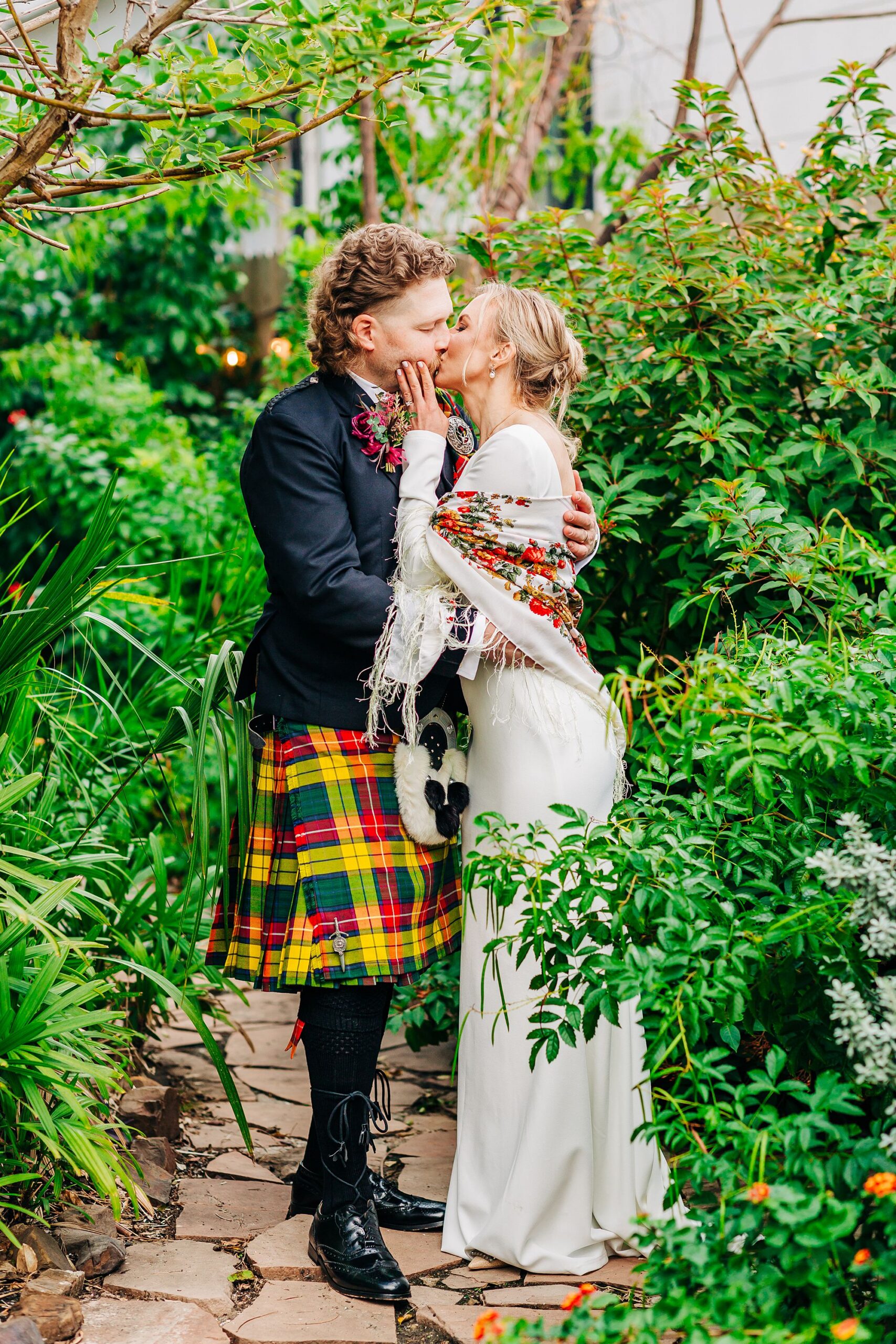 A bride in a white dress and floral shawl and a groom in kilt kiss in a garden surrounded by greenery.