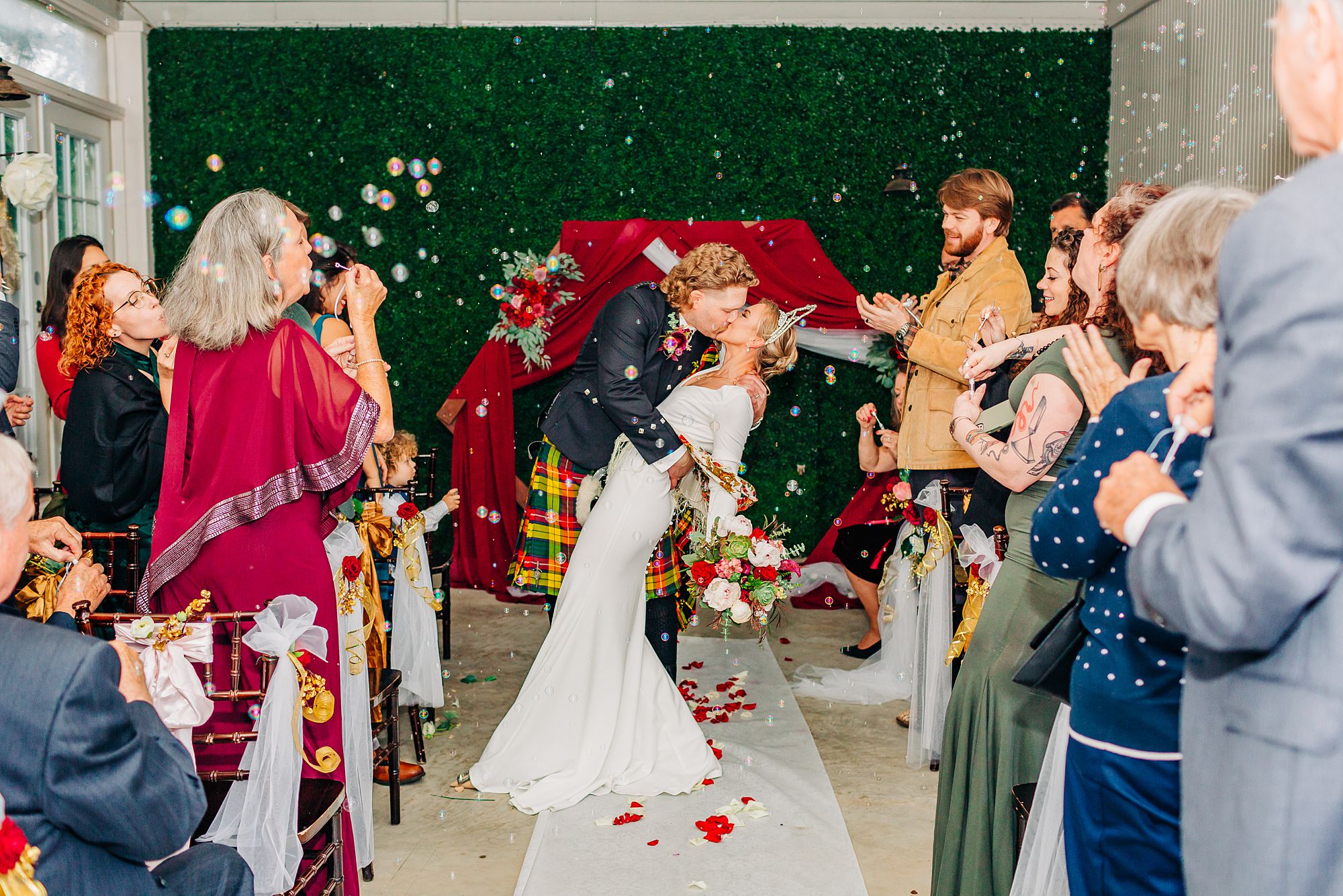 A groom dips and kisses his bride on their ceremony aisle as guests blow bubbles all around them.