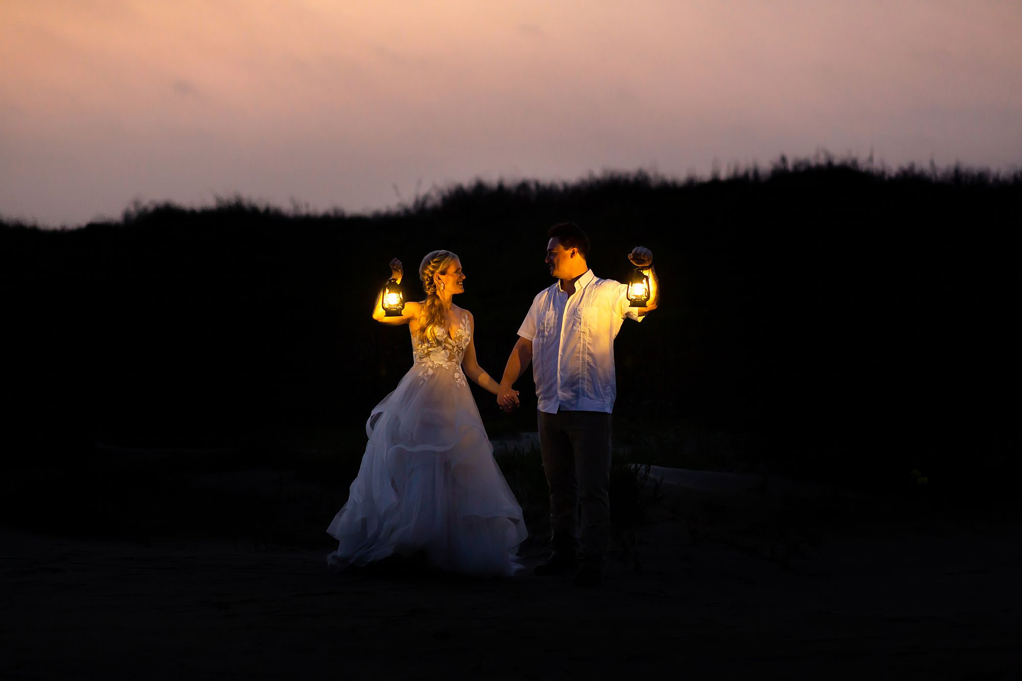 A bride and groom holding lanterns stand in front of a sand dune with a pink sunrise behind it. The couple is looking at each other and smiling.
