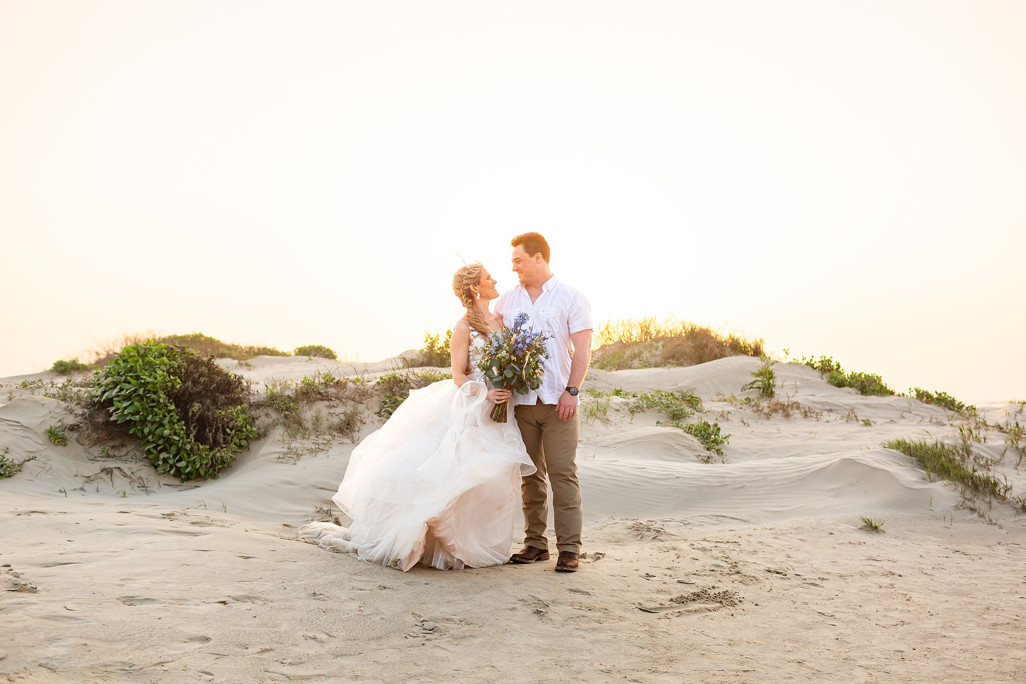 A bride and groom stand near a sand dune with the glow of the sunrise behind them.