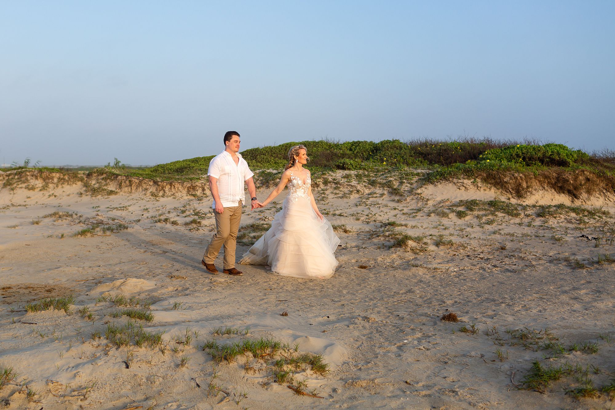 A bride and groom walk past a sand dune with green vegetation, at sunrise on Galveston Island's east end.