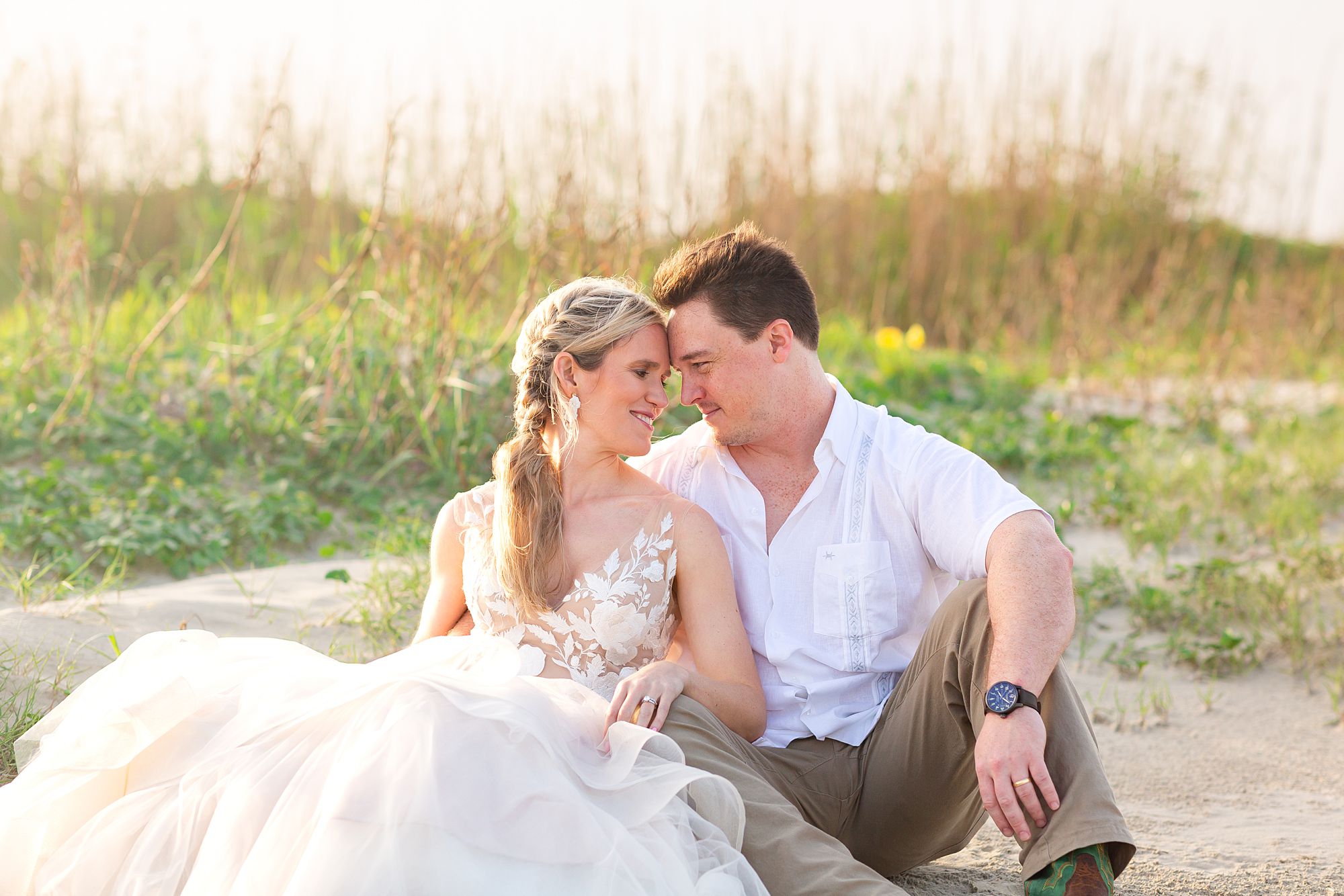 A bride and groom sit nestled together in the sand near a dune with the glow of the sunrise behind them.