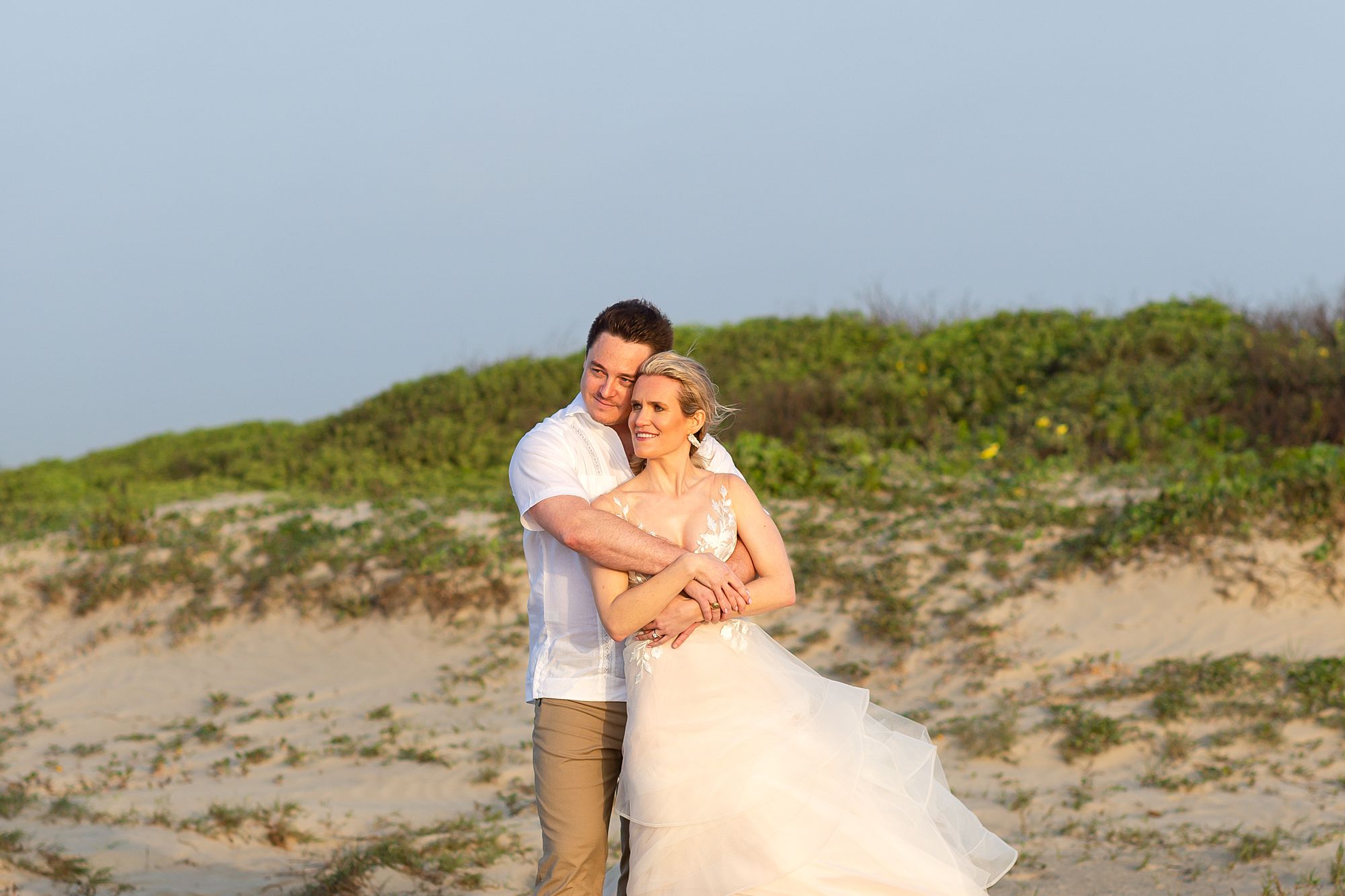 A groom embraces his bride on a Galveston beach during sunrise. Behind them is a sand dune with green vegetation and yellow flowers.