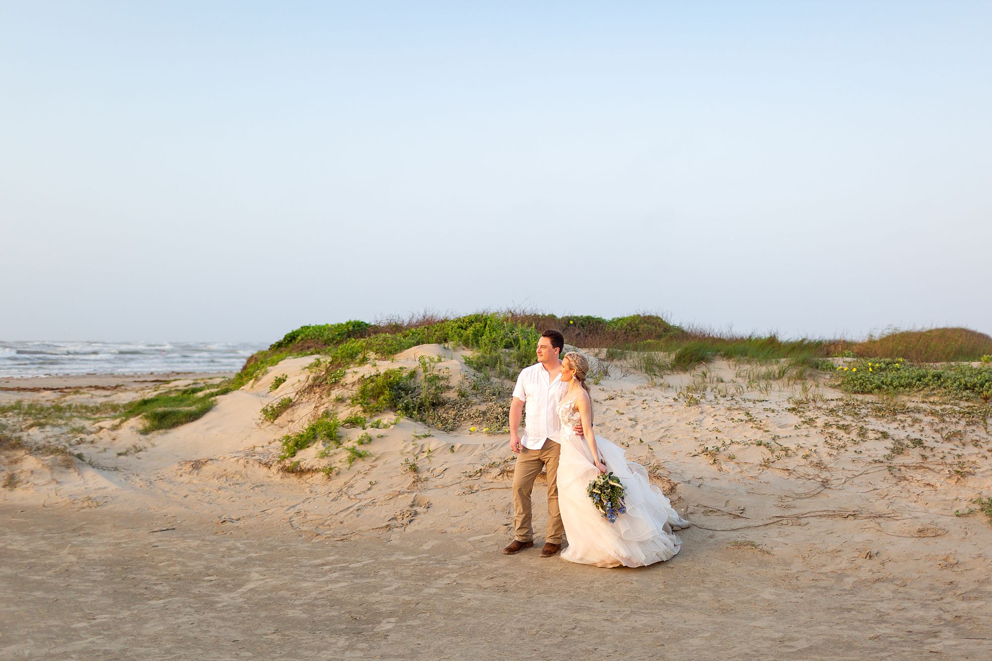 A bride and groom stand in front of a sand dune looking out at the Gulf of America after their sunrise Galveston beach elopement ceremony.