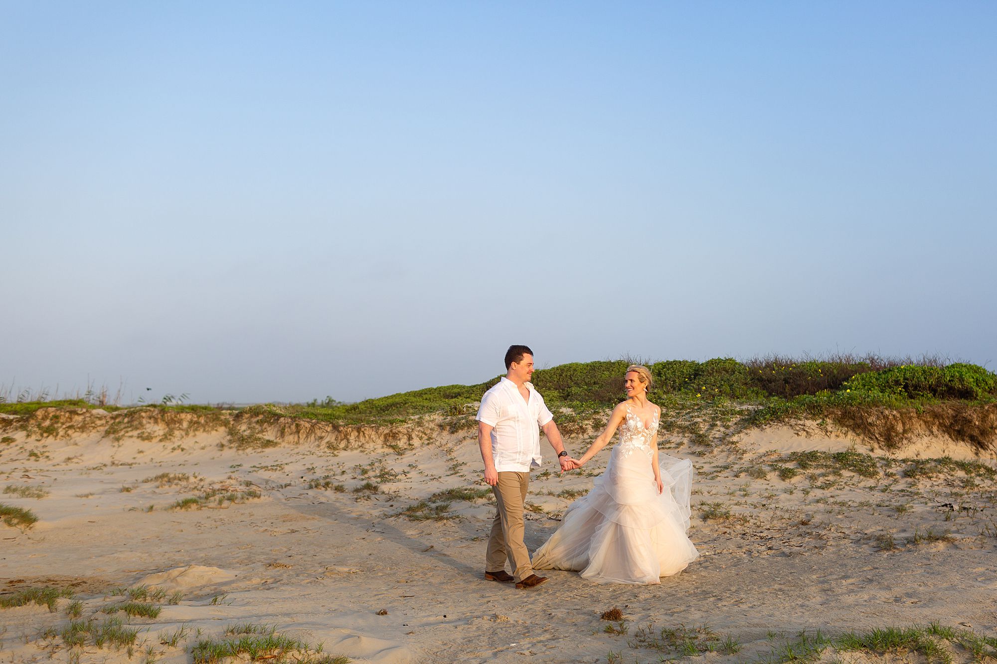 A bride in a tulle ballgown leads her groom along the beach on Galveston Island during their sunrise elopement.