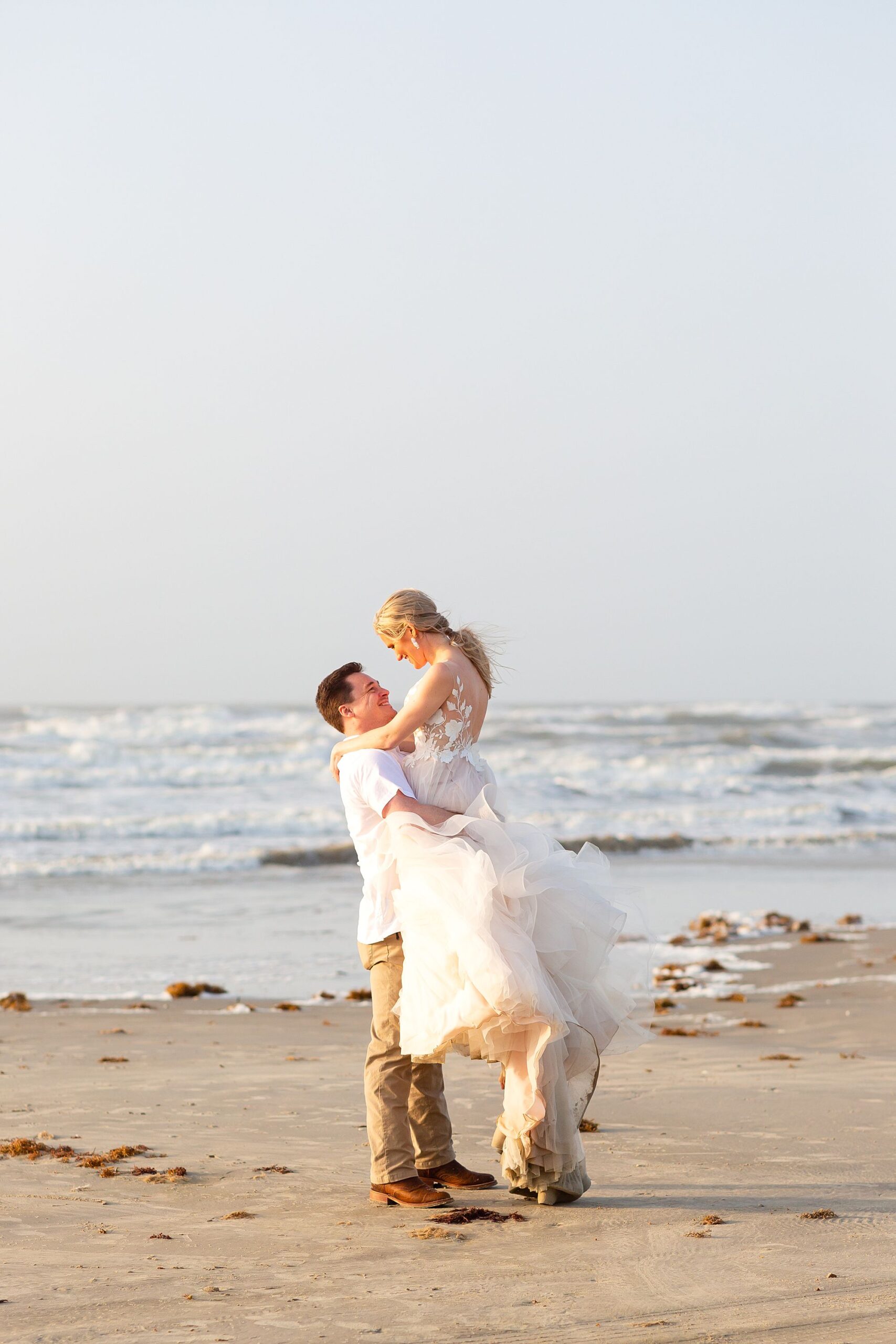 A groom lifts his bride on the beach with the Gulf of America in the background. The bride wears and tulle ballgown and smiles at her groom.