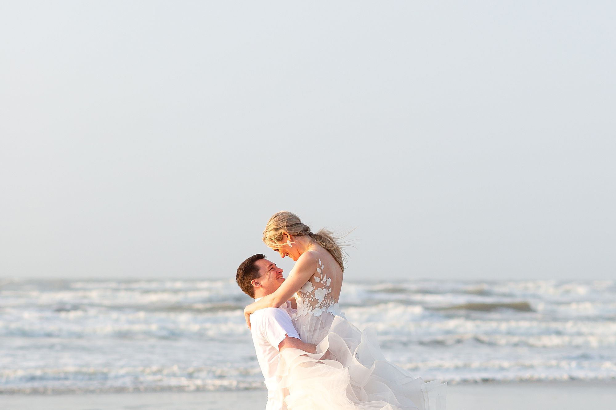A groom lifts a bride up in the air during their sunrise Galveston beach elopement.