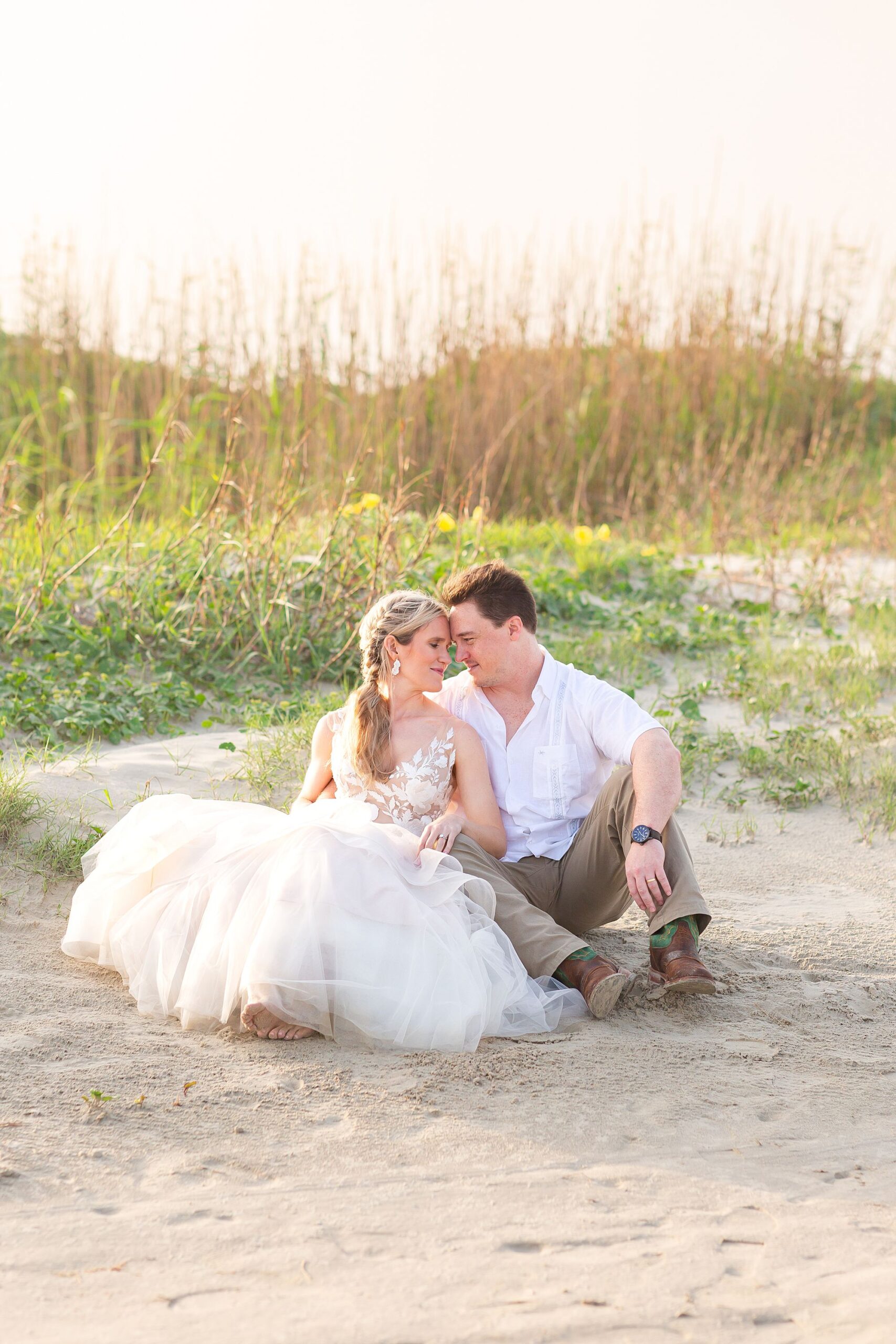 A bride in a tulle ballgown and a groom sit in the sand resting their heads together during sunrise in Galveston. A dune with green vegetation and yellow flowers is behind them.