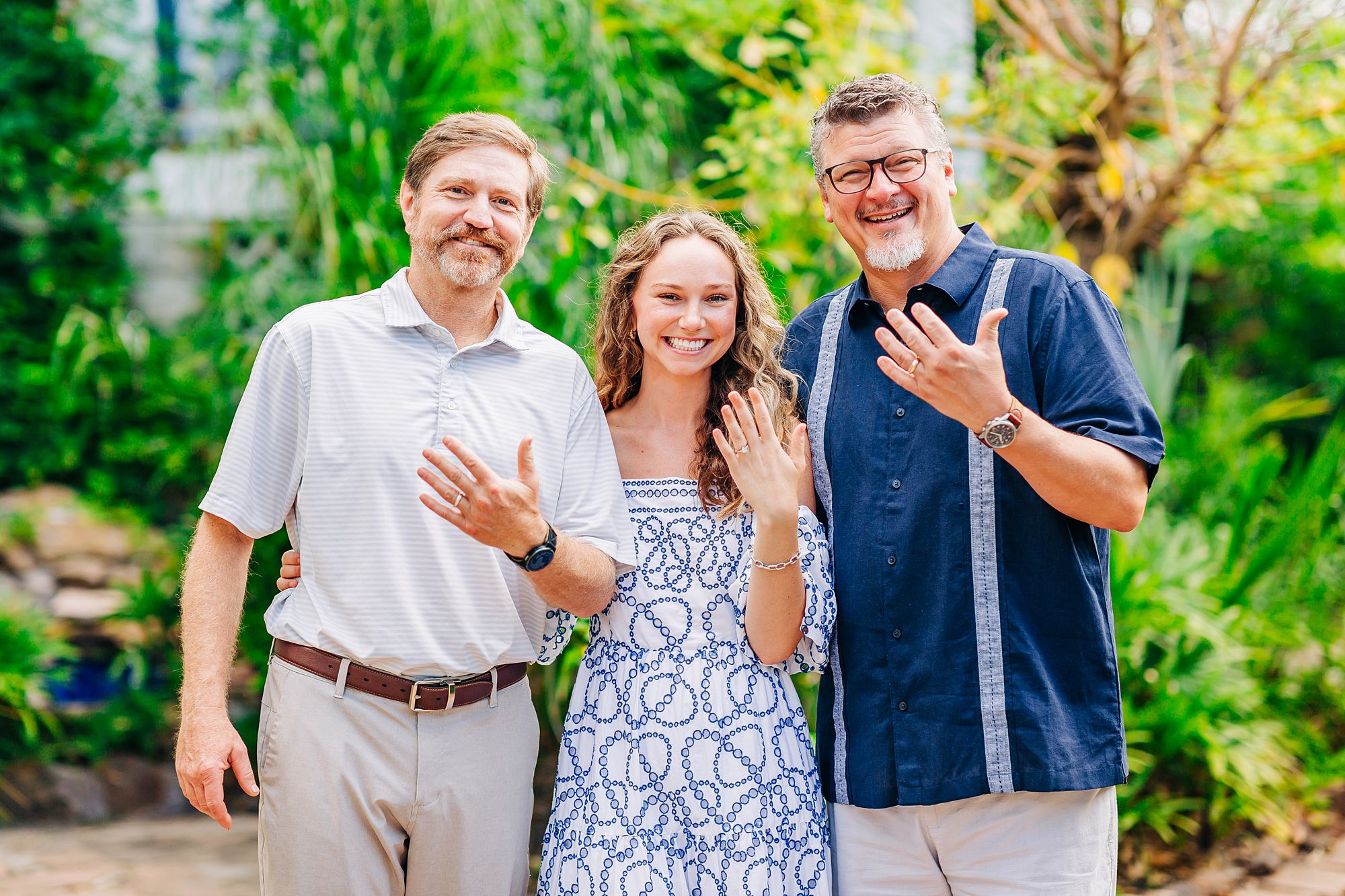 A bride to be poses with her dad and future father in law showing off their engagement and wedding rings.